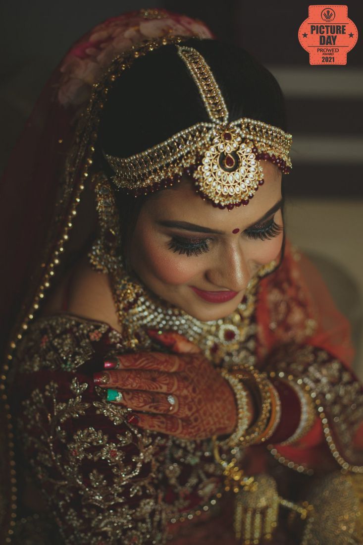 Indian bride in red lehenga with gold maang tikka, nath, and layered jewelry, hands adorned with mehndi and rings.