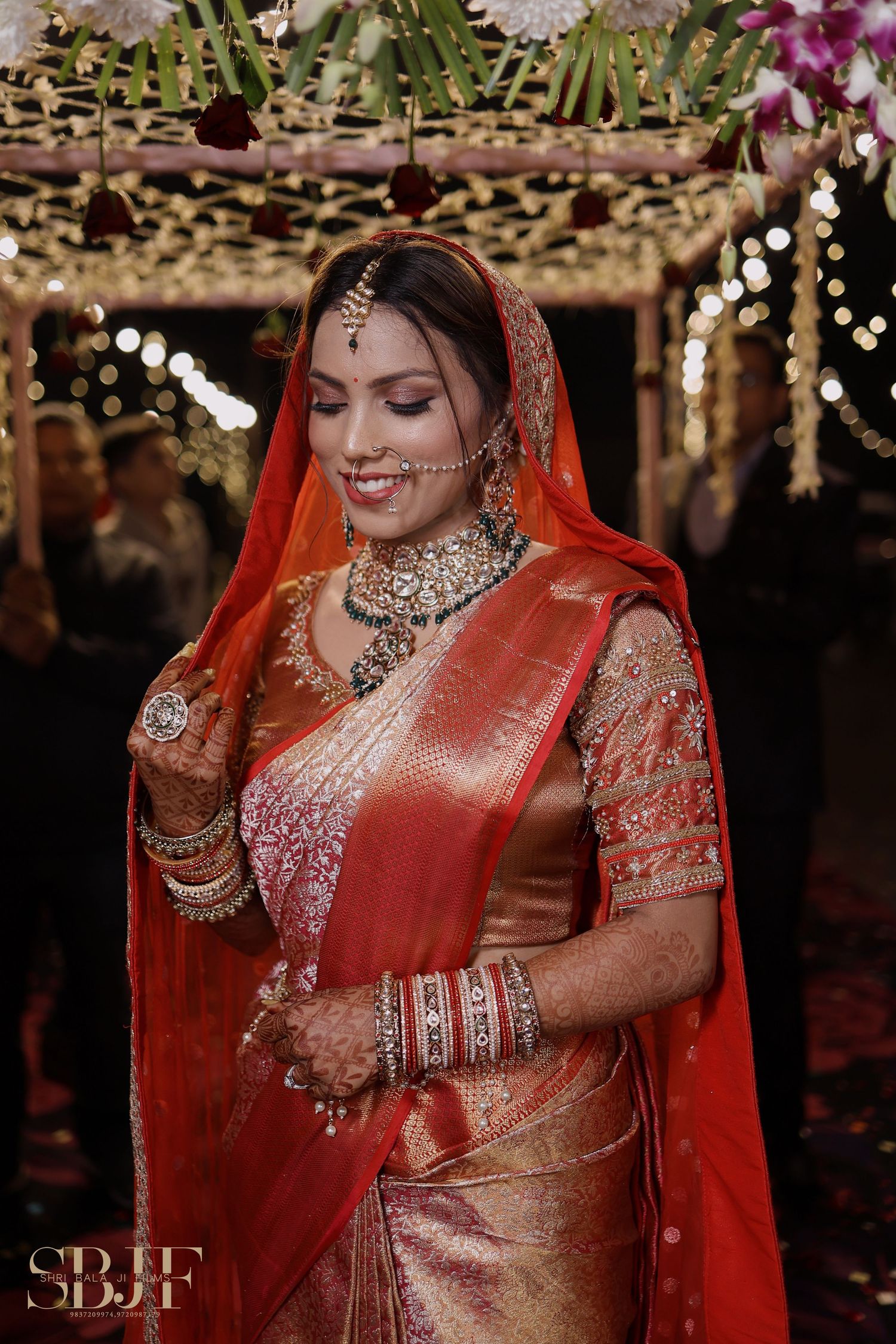 Traditional Indian bridal attire featuring a red and gold silk saree with ornate jewelry under decorative lighting at a wedding.