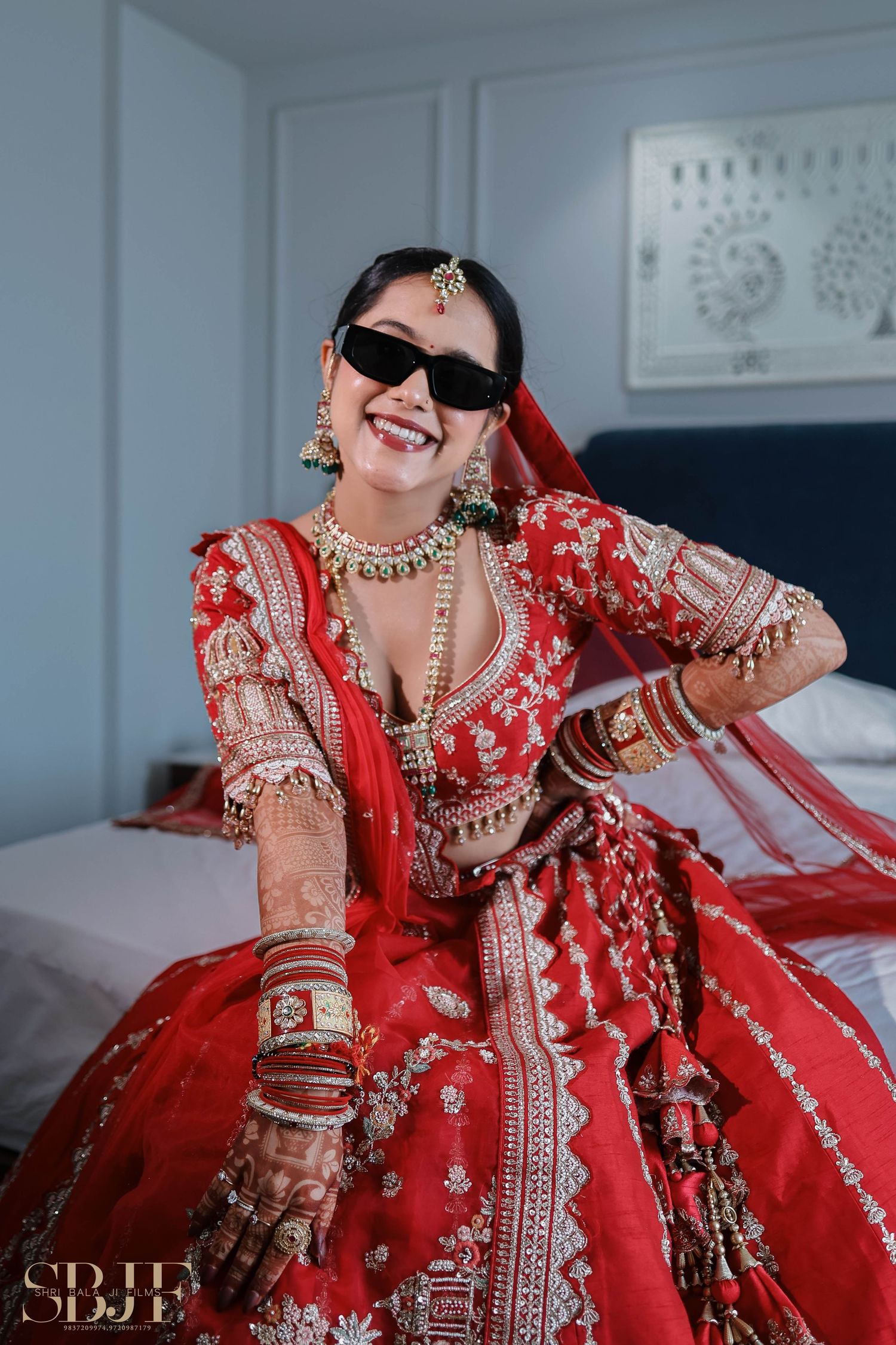 Traditional Indian bride wearing an ornate red lehenga with detailed embroidery and jewelry sits on white bedding.