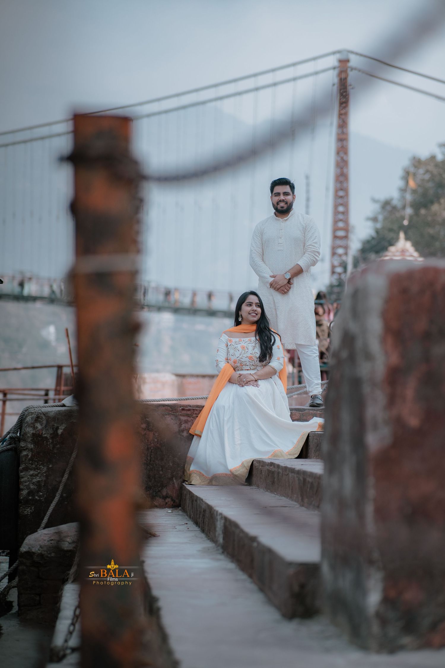 Couple in formal attire pose on concrete steps with bridge cables visible in background.