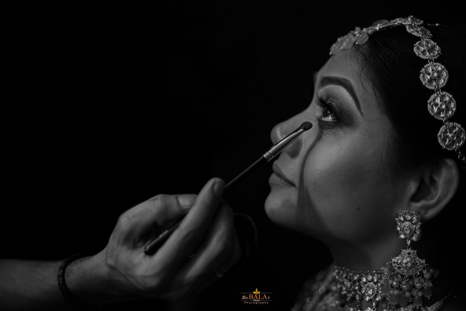 Close-up black and white photograph of makeup being applied to a bride wearing traditional jewelry.