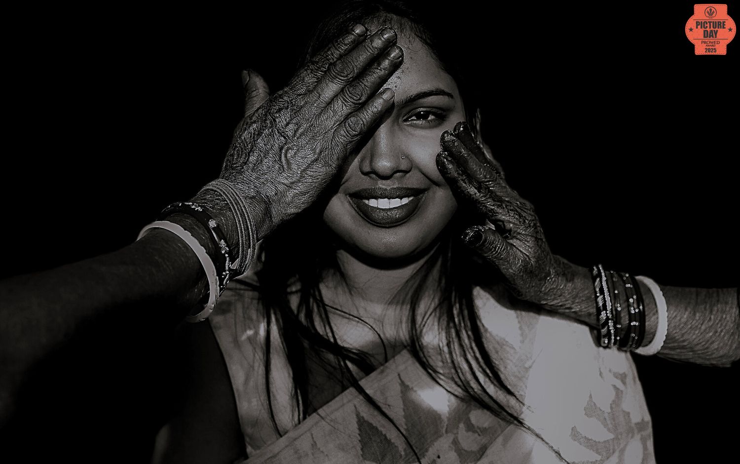 Black and white photo of a smiling Indian woman with henna-decorated hands covering her face.