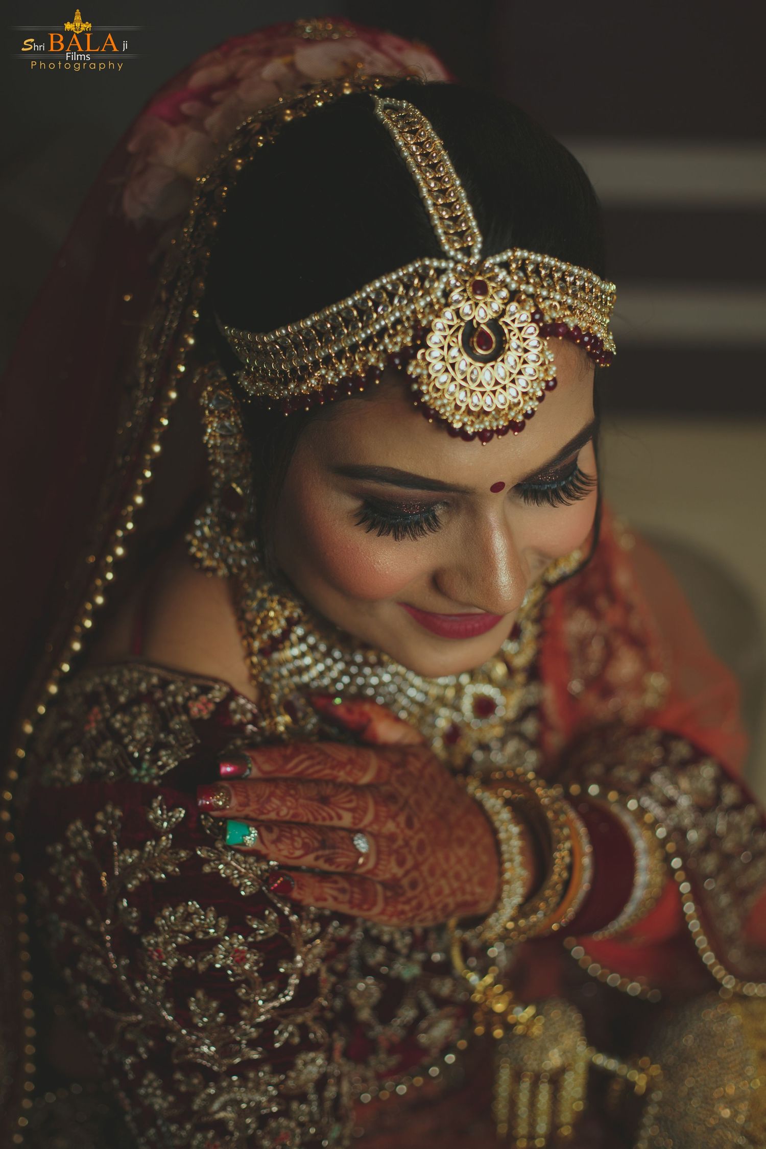 Traditional Indian bride wearing ornate gold jewelry and red bridal attire with mehndi adorned hands looking down demurely.
