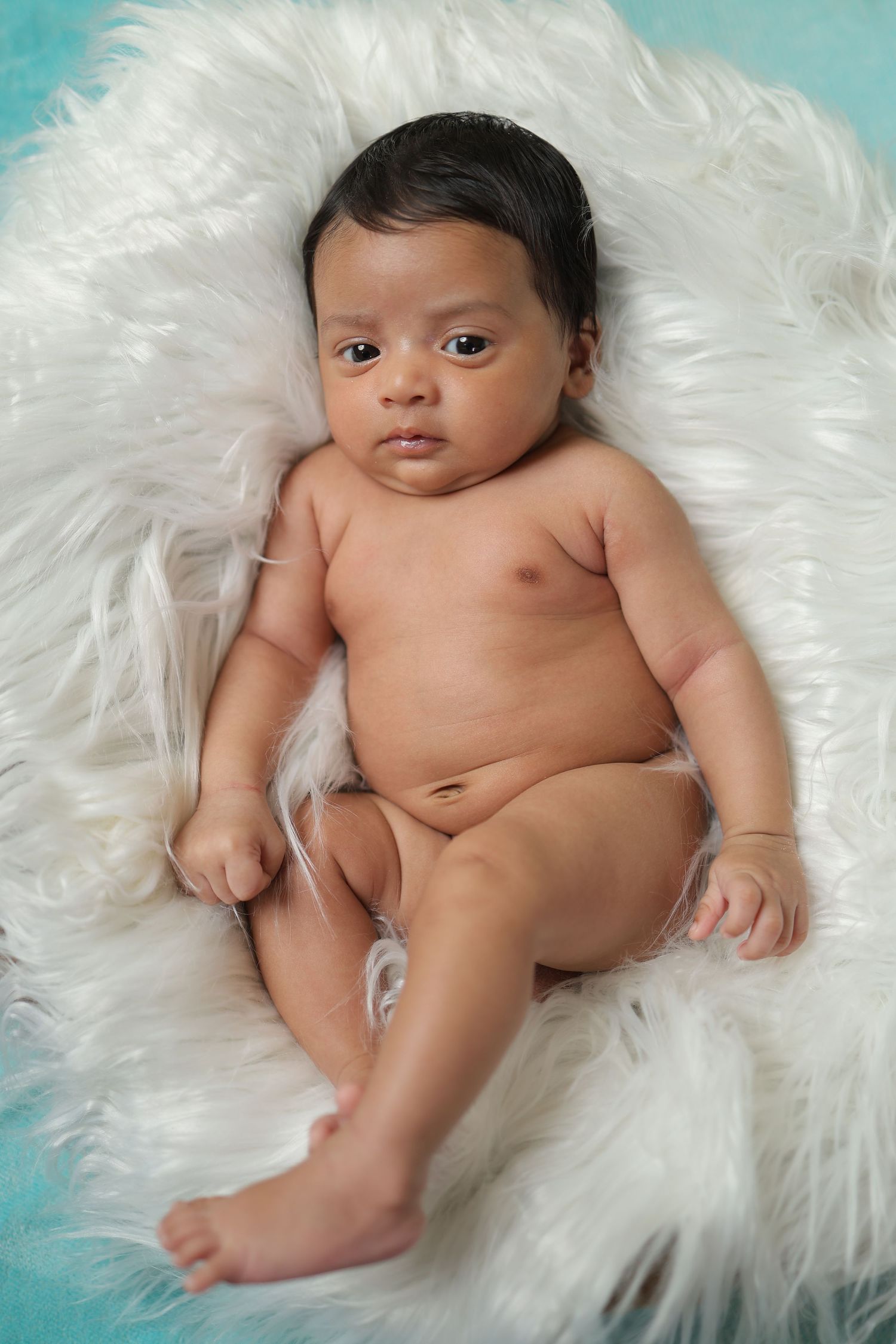 Newborn baby lying on soft white fur blanket against turquoise backdrop in professional photo session.