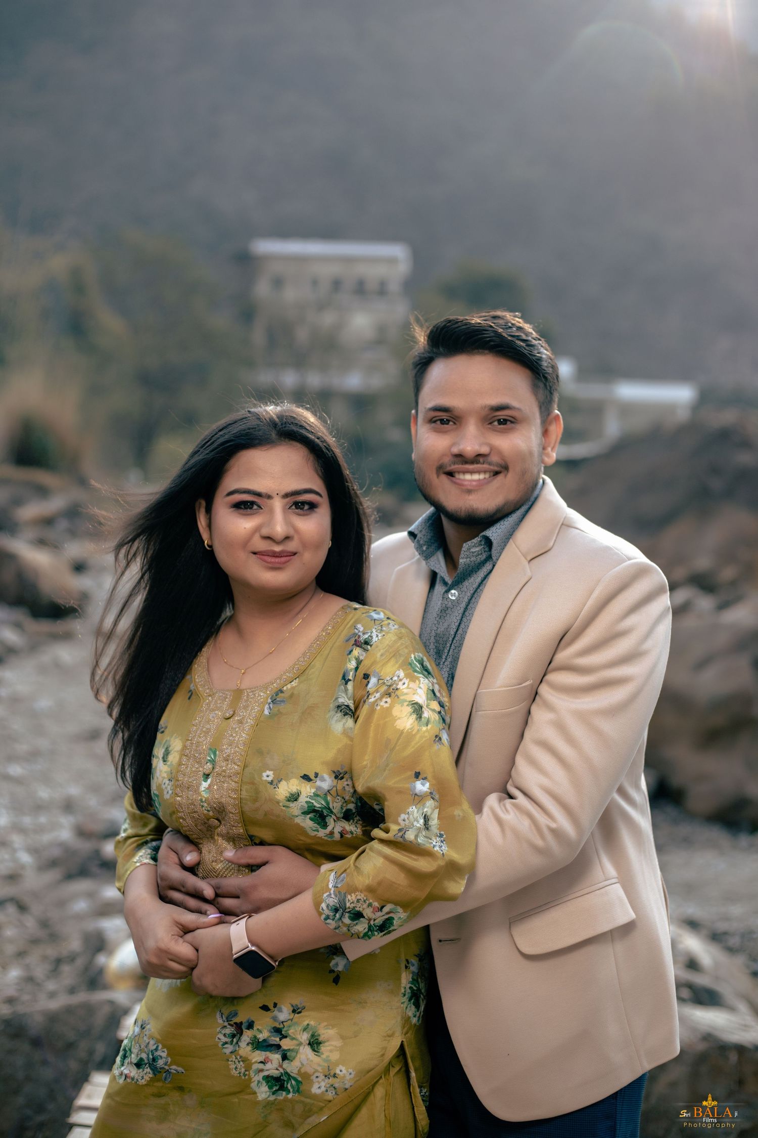 Couple in casual formal wear posing against mountainous landscape.
