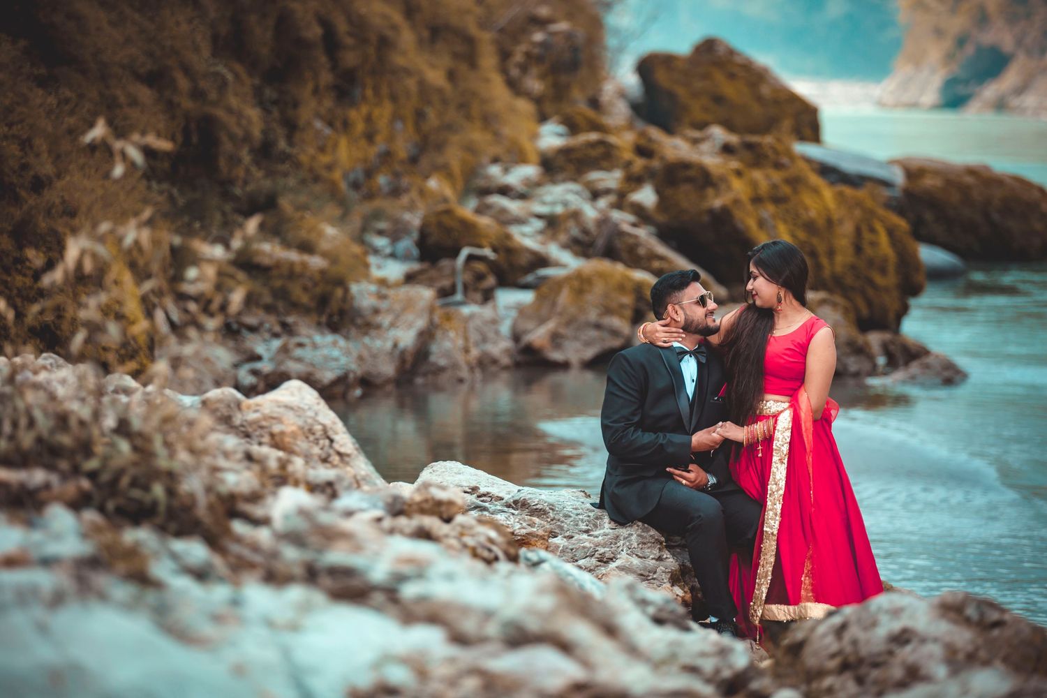 Couple in formal attire sharing a romantic moment on rocky coastal terrain.