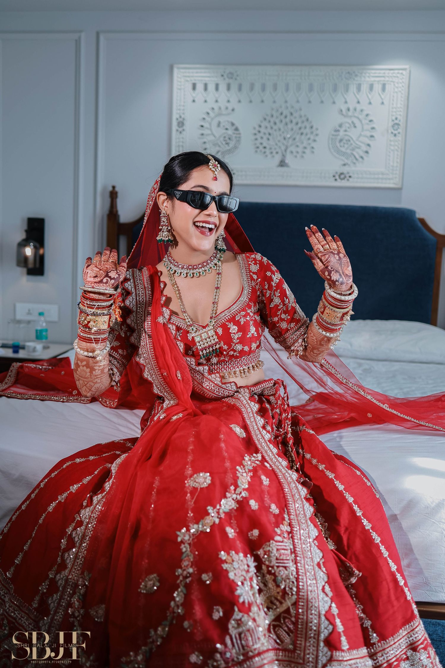 A bride in a traditional red lehenga and sunglasses sits on a bed wearing ornate jewelry and henna designs.