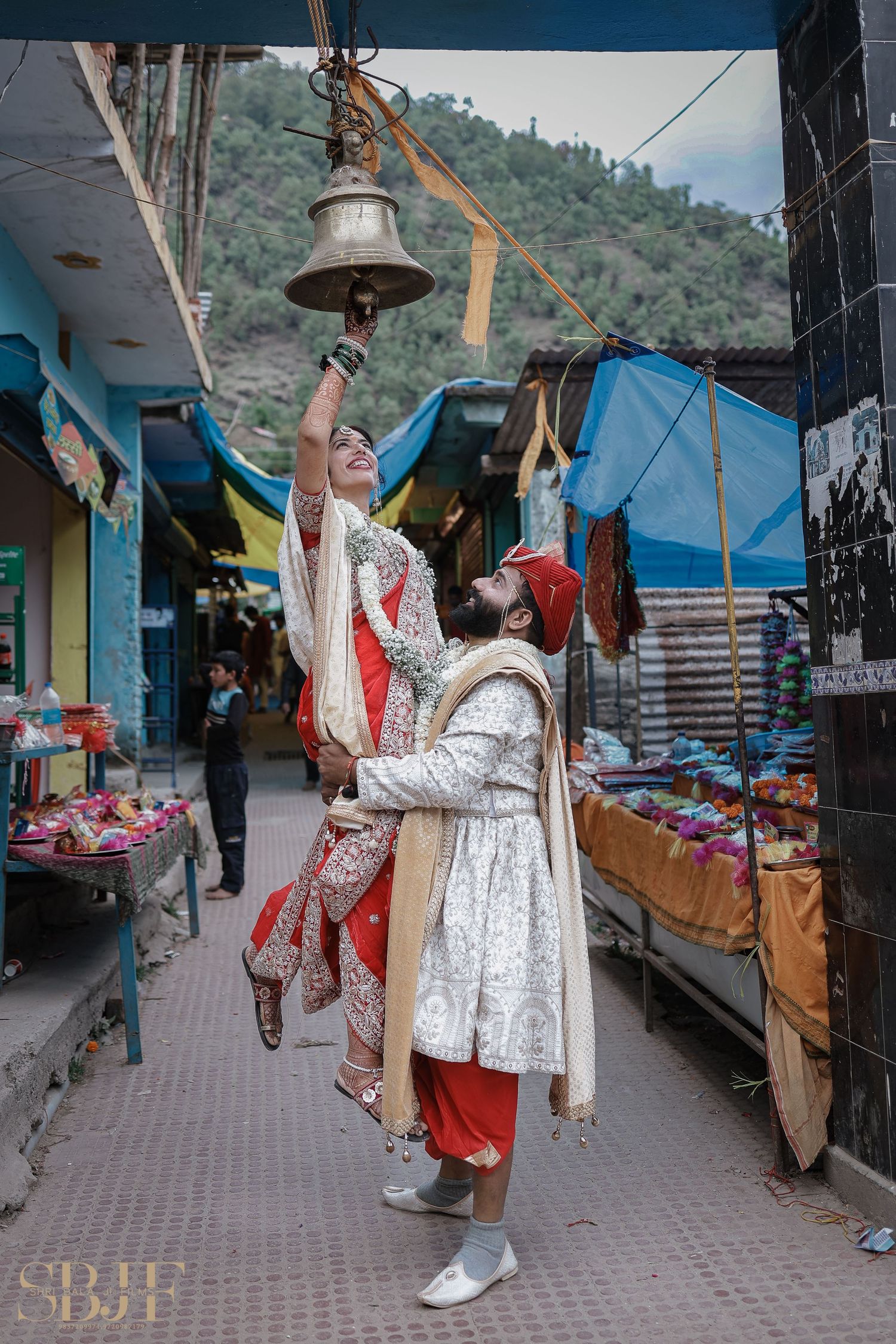 Indian bride and groom in traditional attire ringing a temple bell together in a vibrant market street.