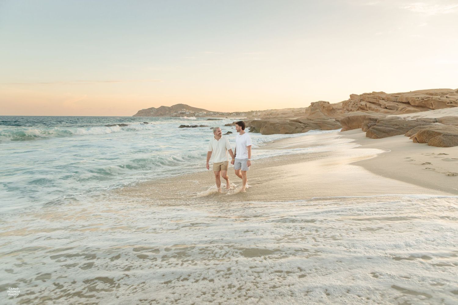Couple walking hand in hand along shoreline with ocean waves and rocky cliffs at sunset.