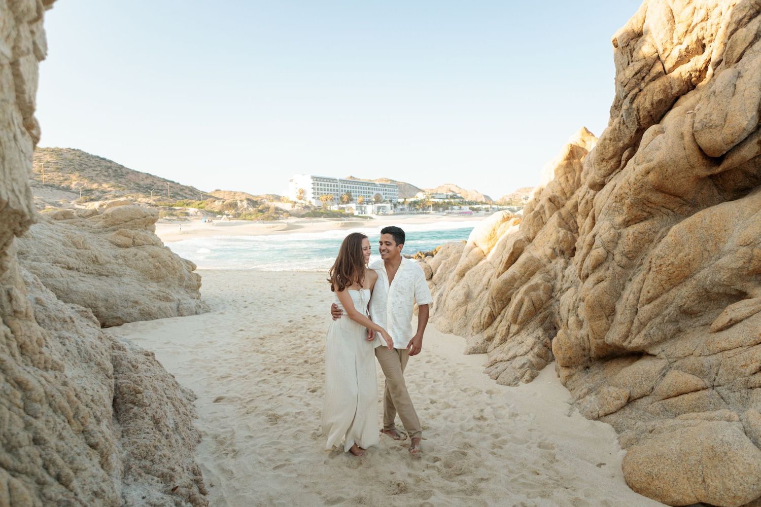 Couple walks hand in hand between white rock formations on a sunny beach.