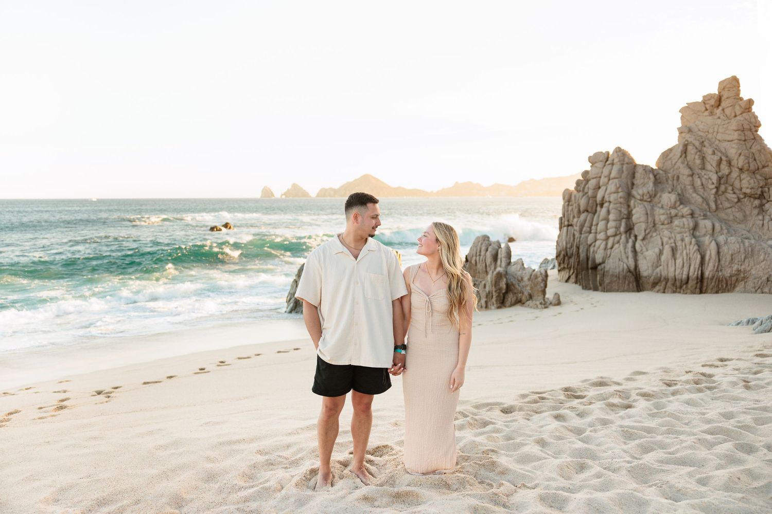 Couple holding hands on a sandy beach with rocky cliffs and ocean waves in the golden hour light.