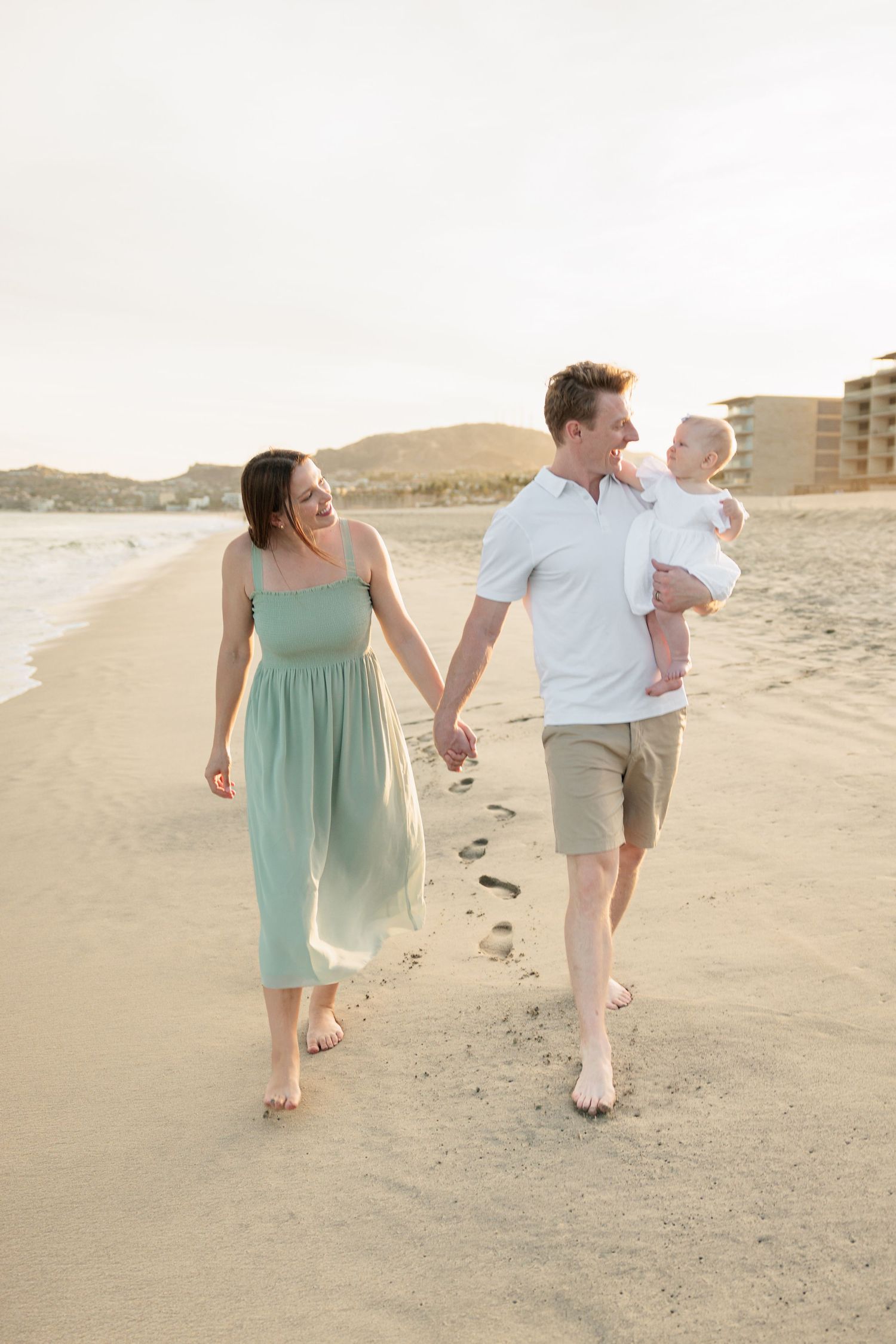 Young family walks barefoot on sandy beach at sunset, parents holding hands while father carries smiling baby.