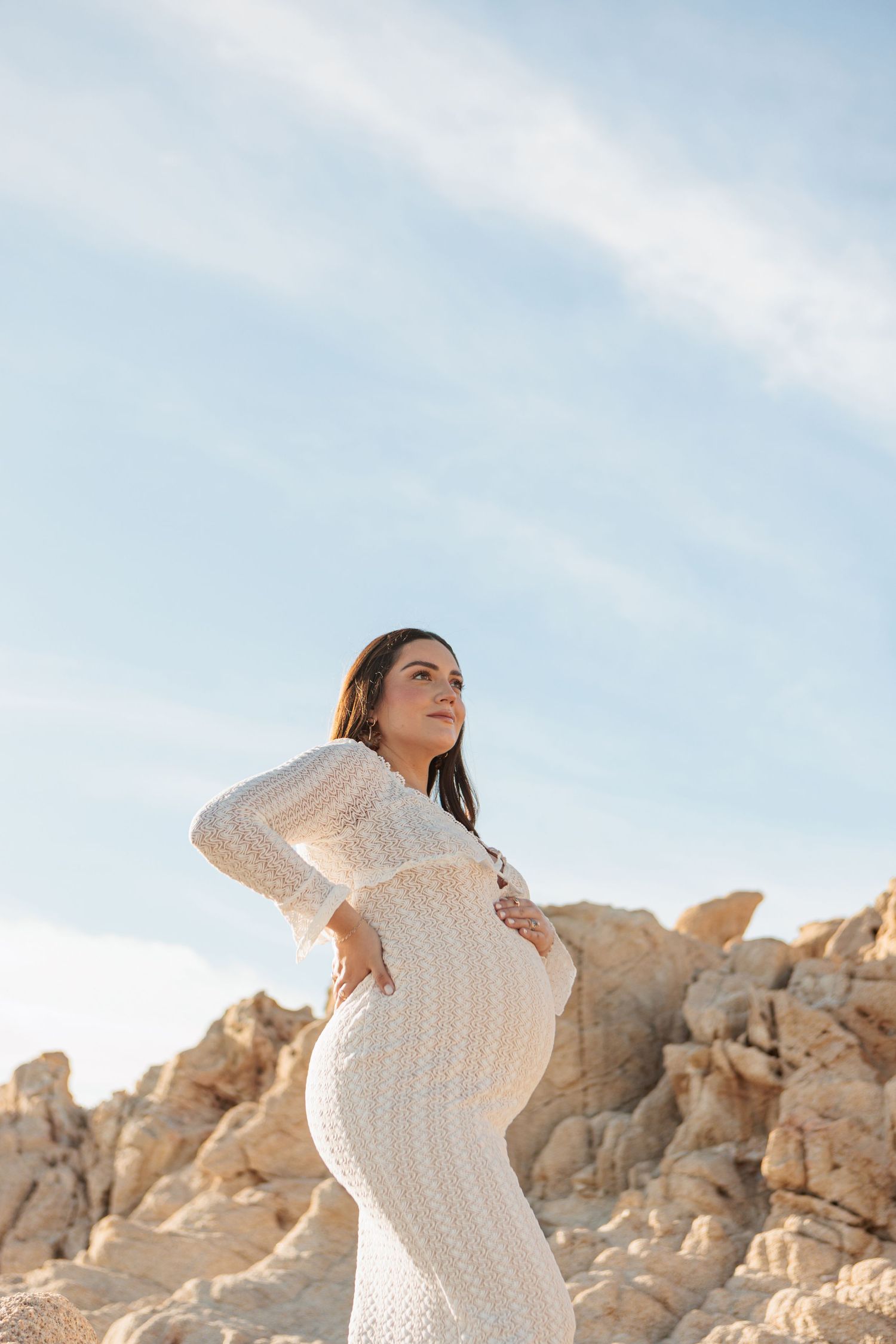 Pregnant woman in white lace dress cradling her baby bump outdoors near rocky terrain under a blue sky.