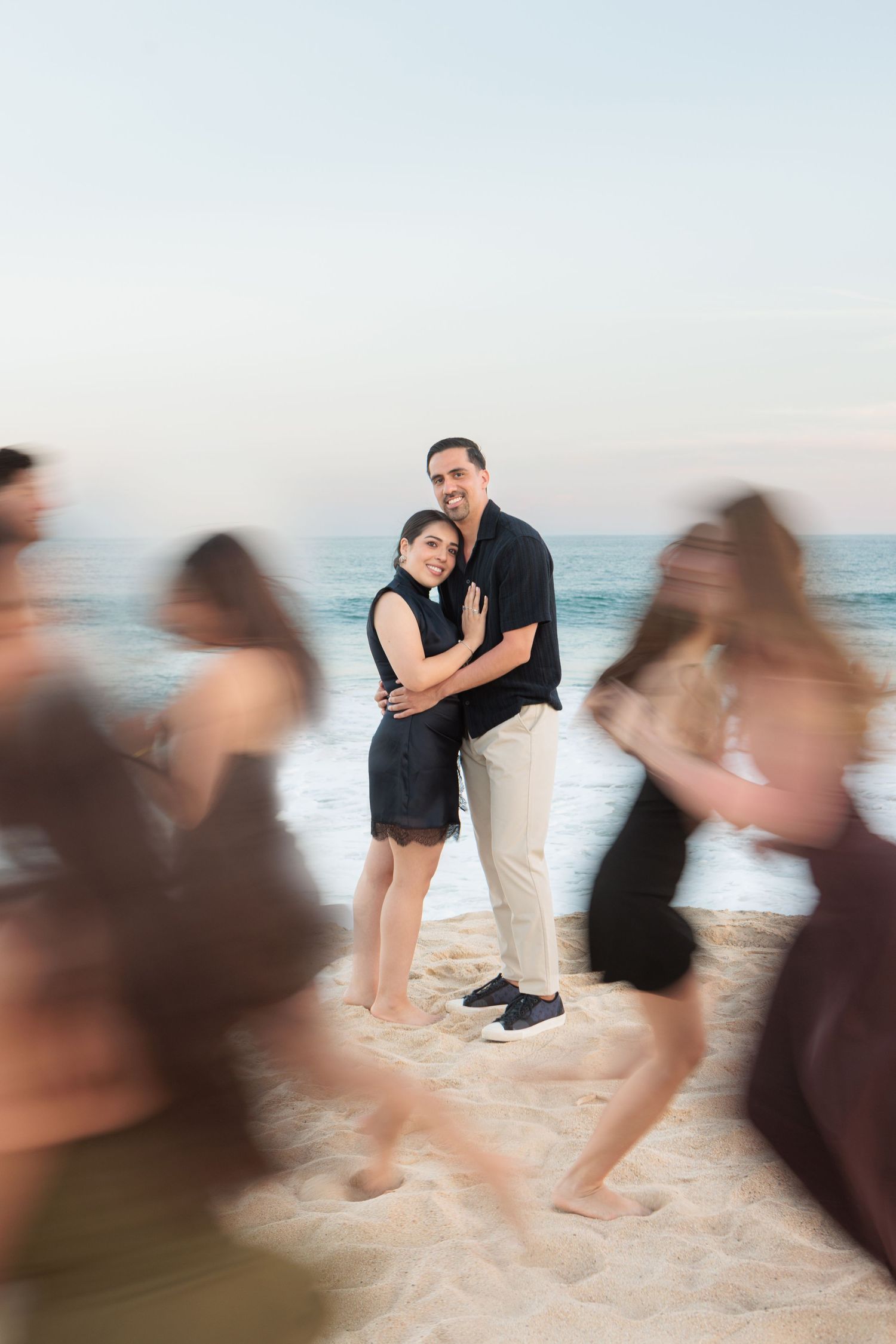 Couple embracing on a sandy beach at sunset, surrounded by blurred moving figures in the foreground.