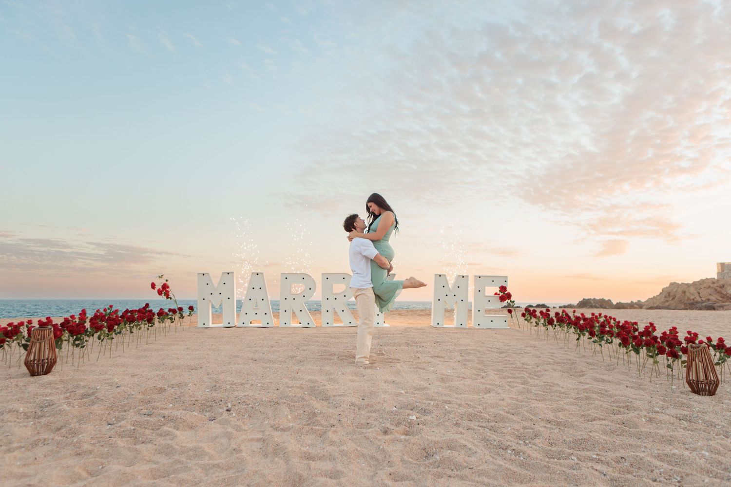 Man lifting woman joyfully at beach proposal with MARRY ME marquee letters and roses.