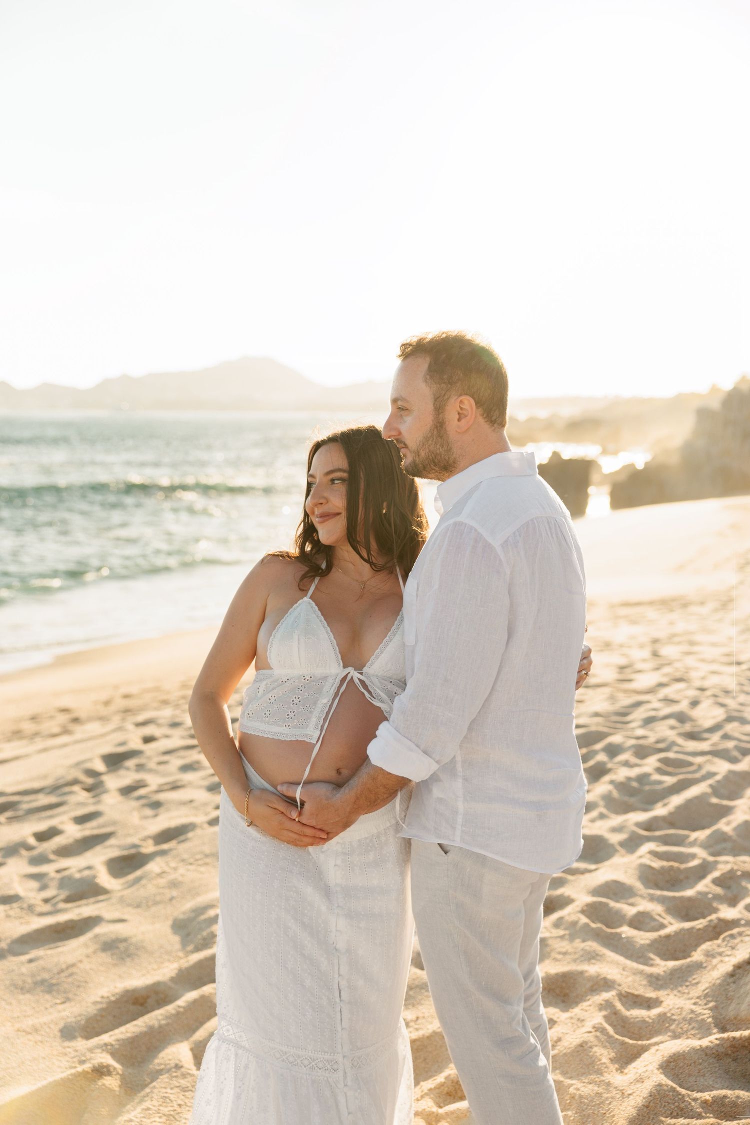 Couple embracing on sunny beach at golden hour, man holding pregnant partners belly tenderly.