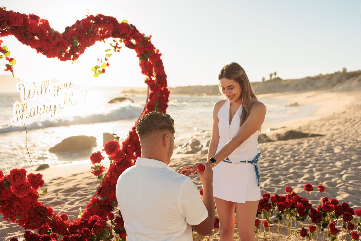 Man proposing to woman in front of a red rose heart arch on a sandy beach.