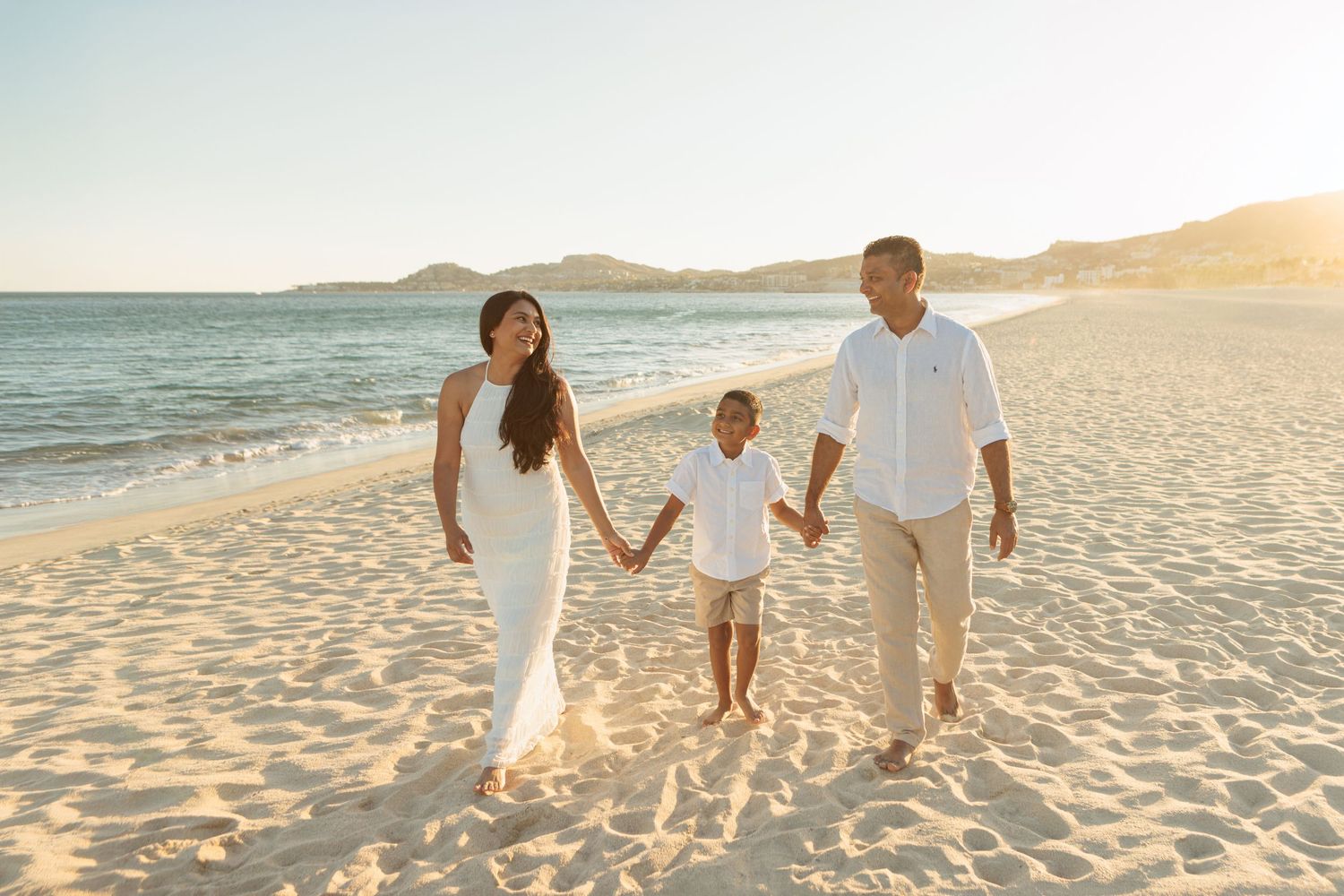Family of three in white outfits holding hands walking along sunny beach with mountains behind.