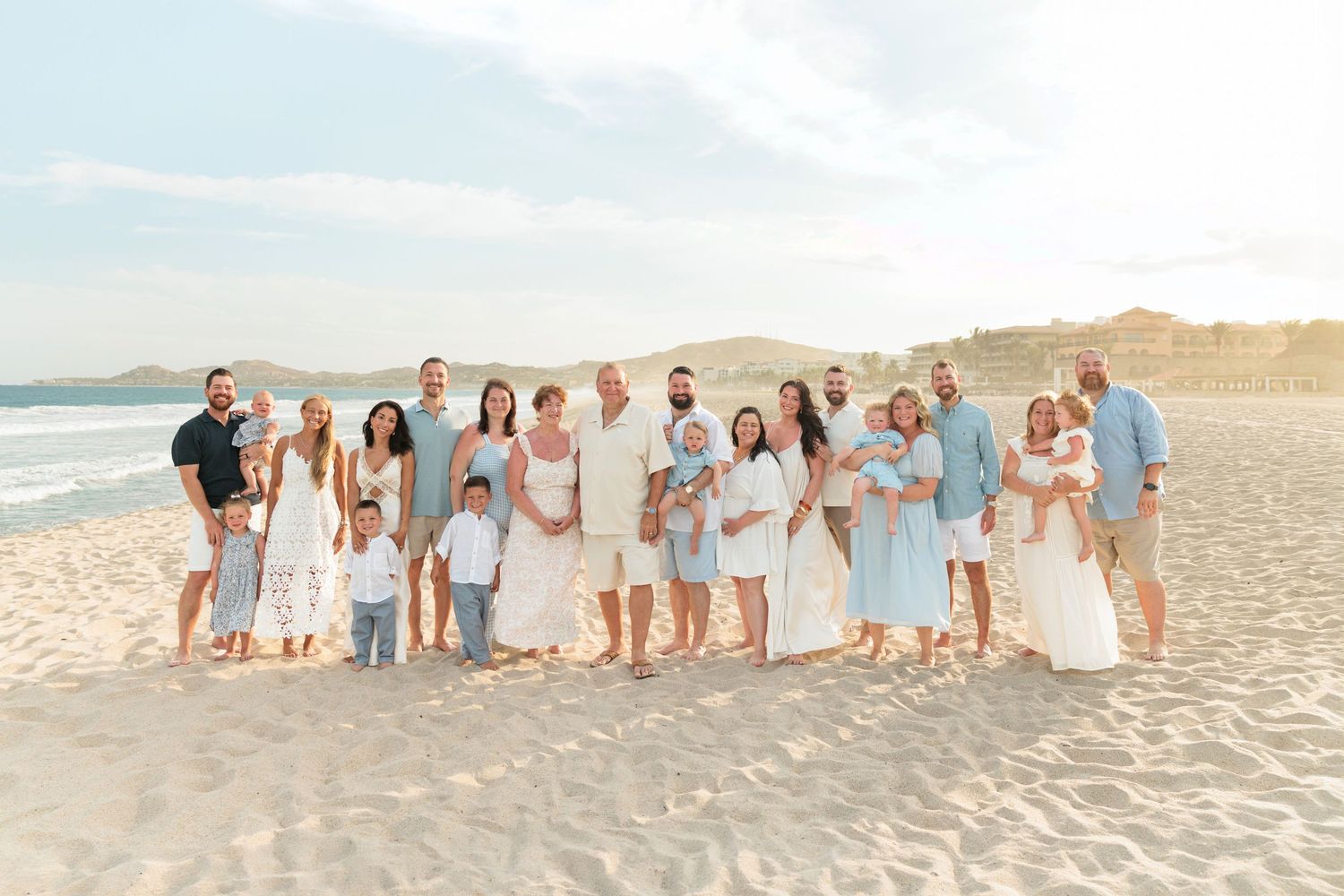 Large family group portrait on a sandy beach at golden hour, dressed in white and light blue outfits.