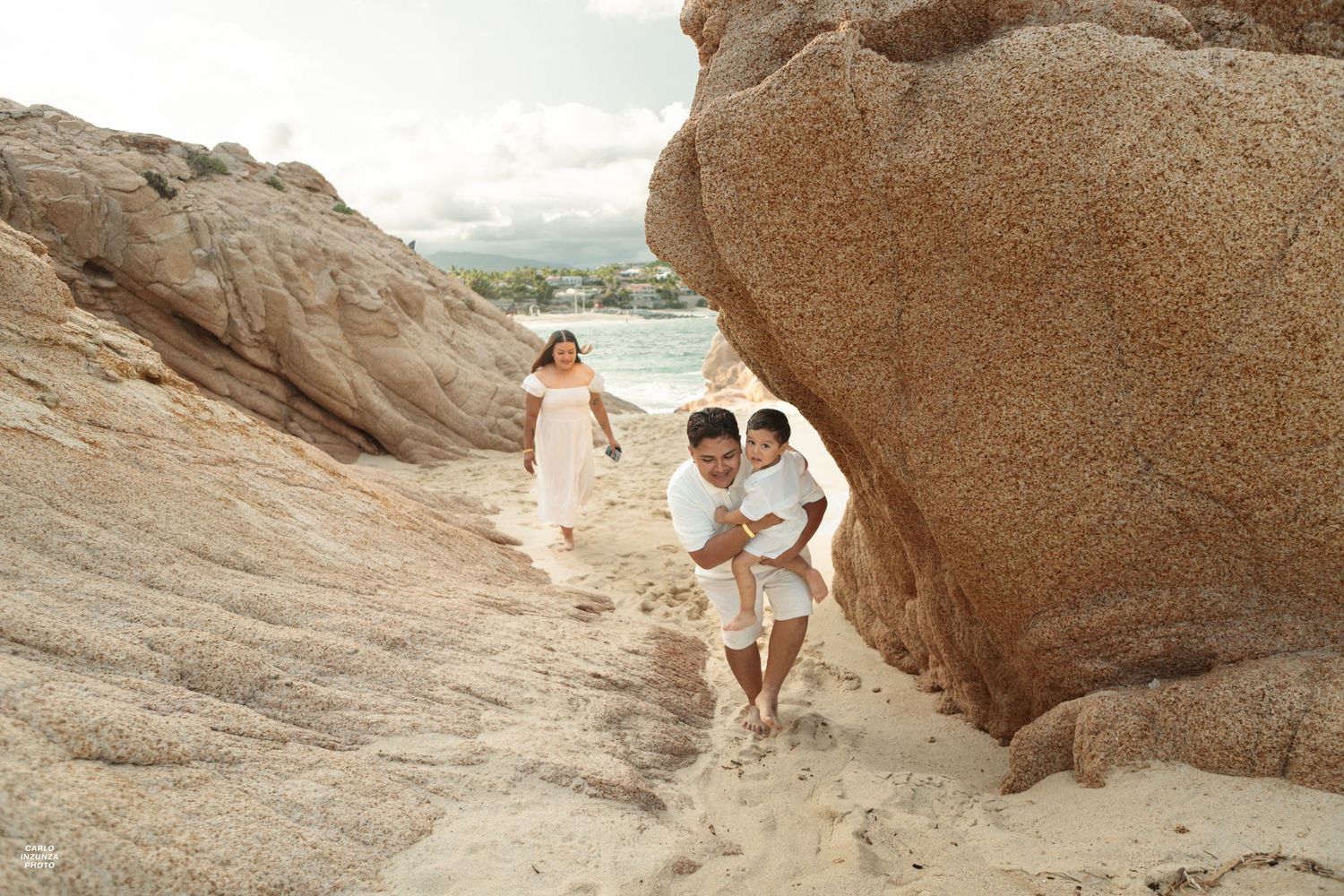 Family in white clothing exploring between large boulders on a tropical sandy beach shoreline.