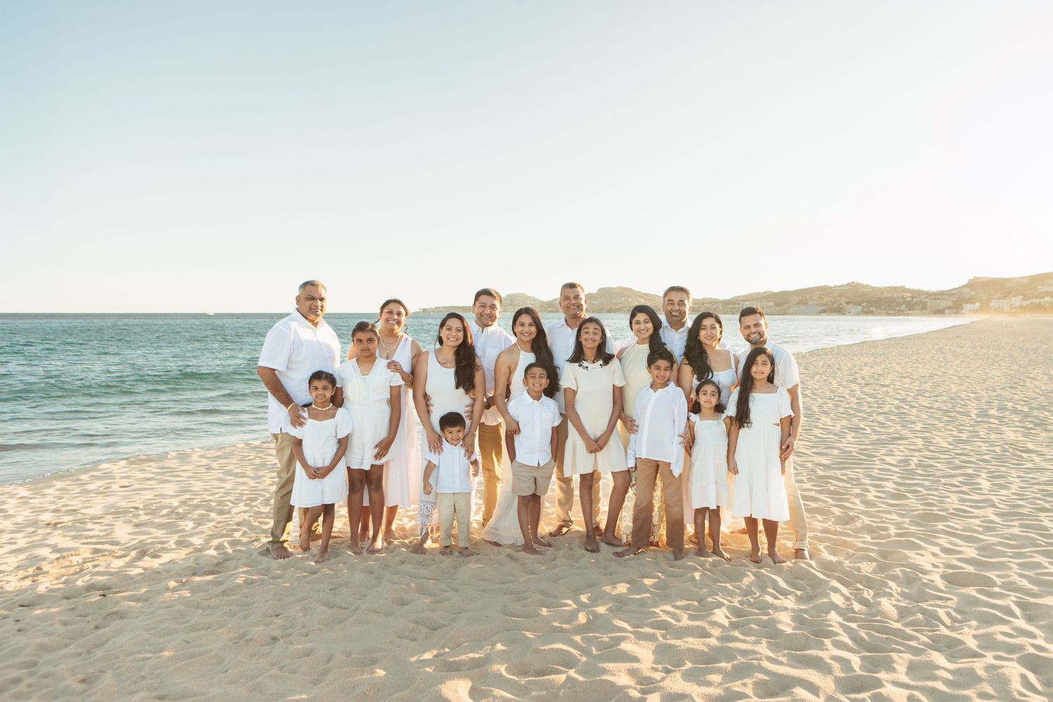 Large family group of adults and children in white outfits posing on sandy beach at sunset.