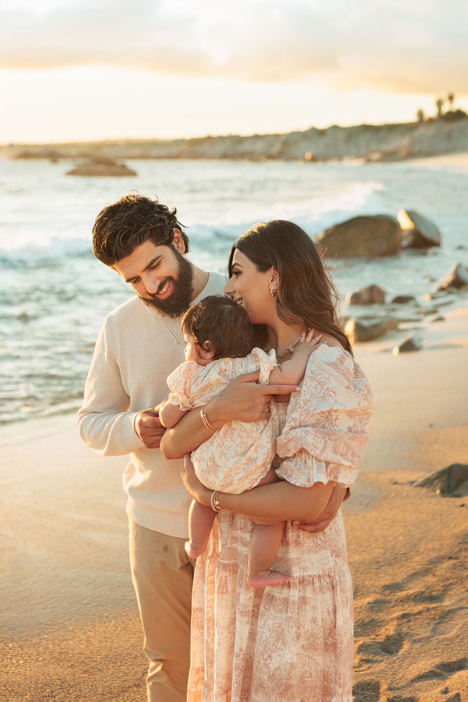 Smiling couple in casual outfits holding their baby girl on rocky beach at golden sunset.