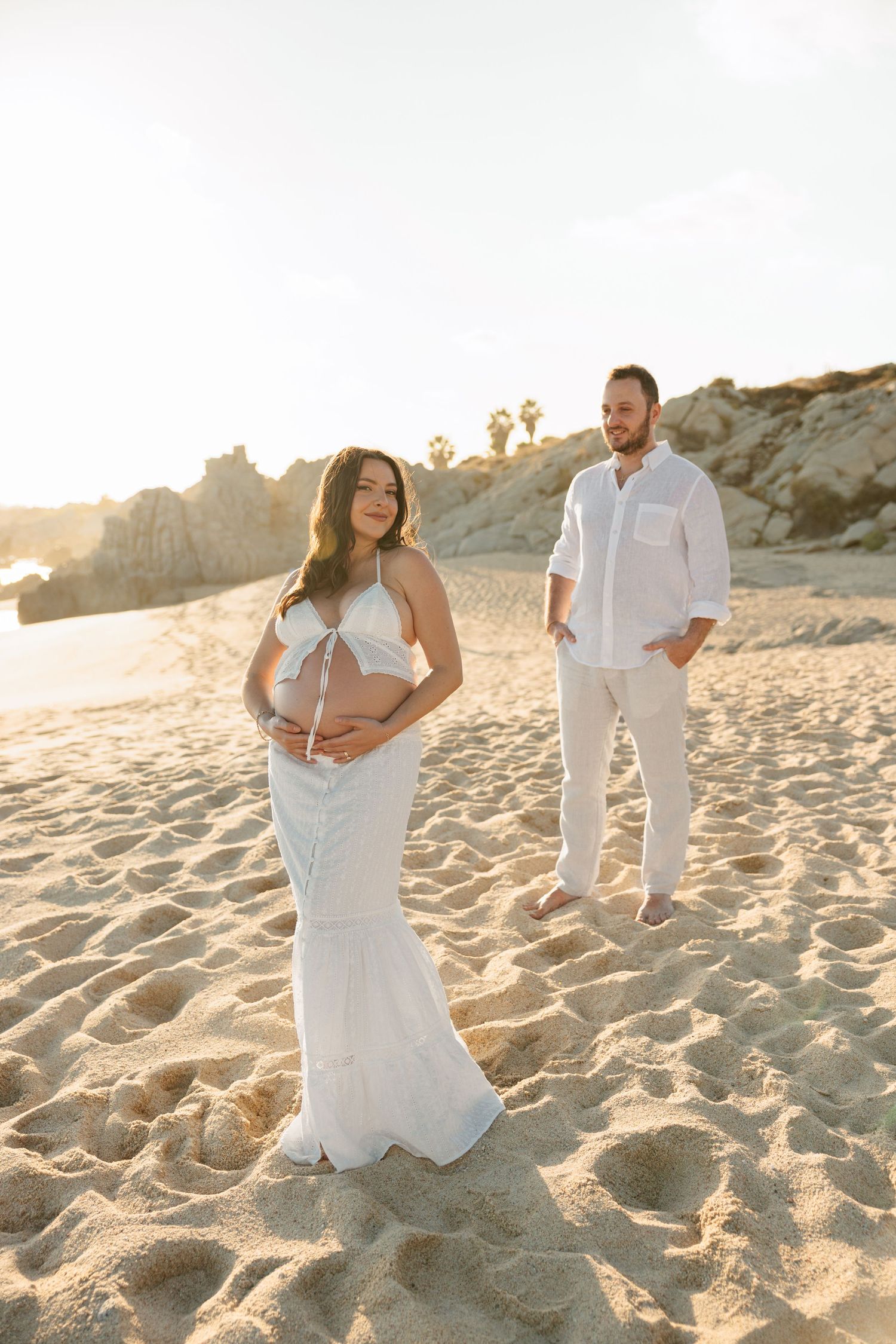 Pregnant woman in white dress holds baby bump on sandy beach at golden hour while man stands behind her.