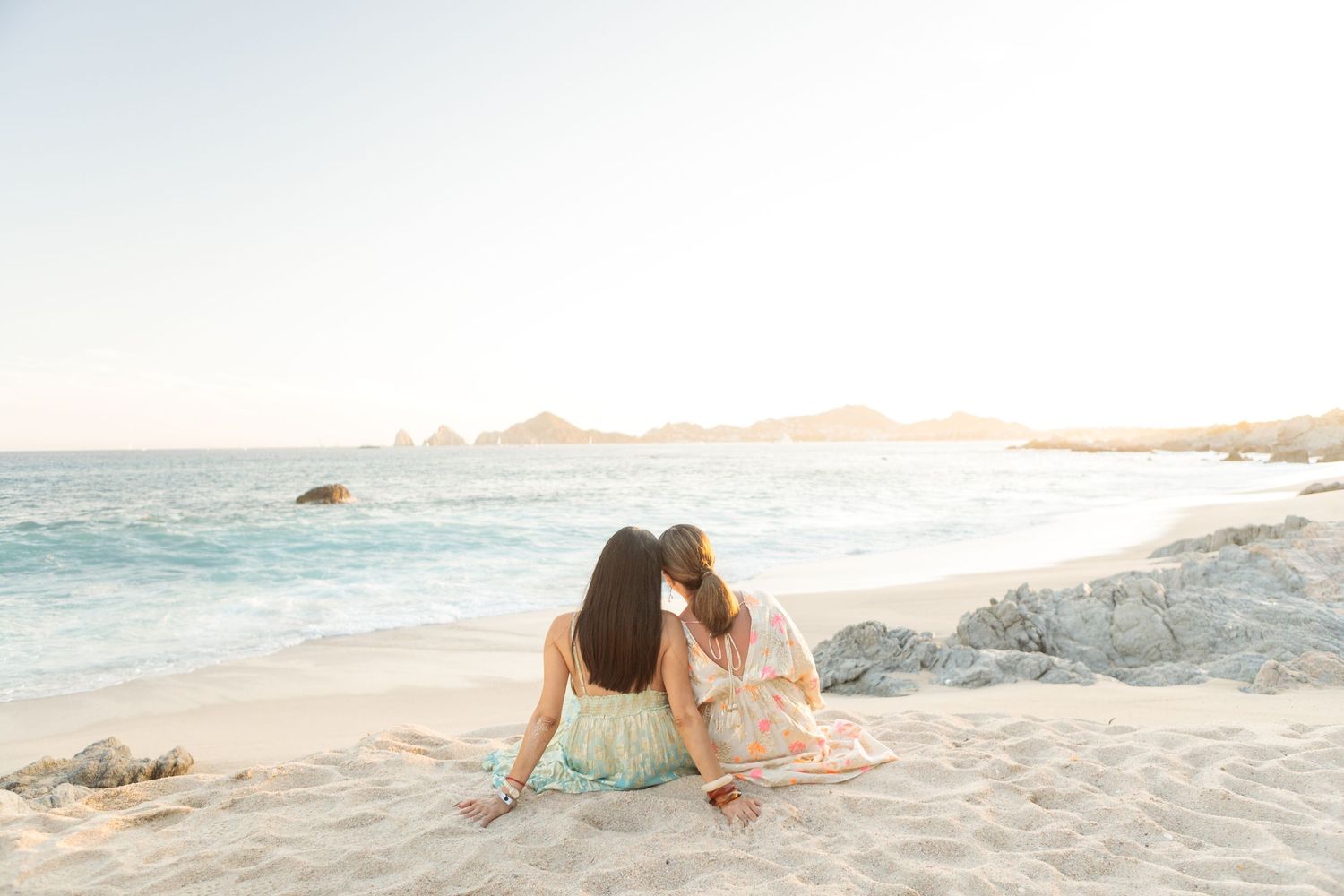 Two women sitting side by side on a beach, facing the ocean with rocky coastline in the background.