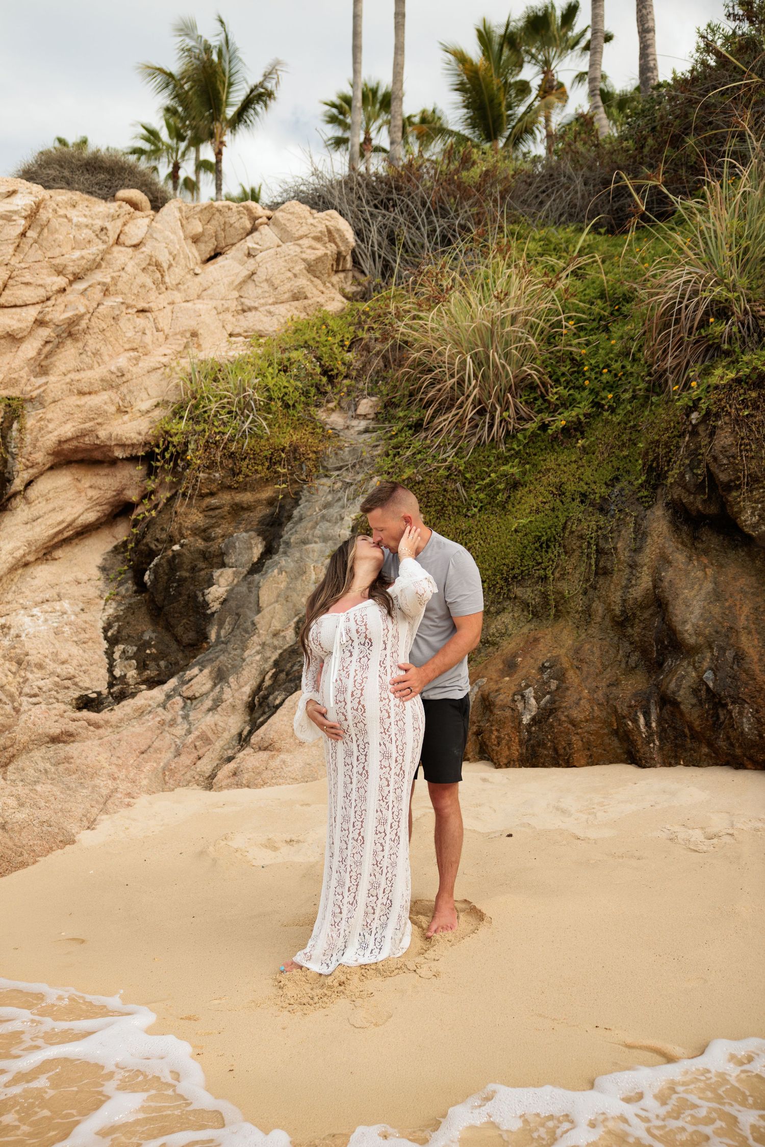 Couple embracing on sandy beach, pregnant woman in white lace dress, rocky cliffs and palm trees backdrop.
