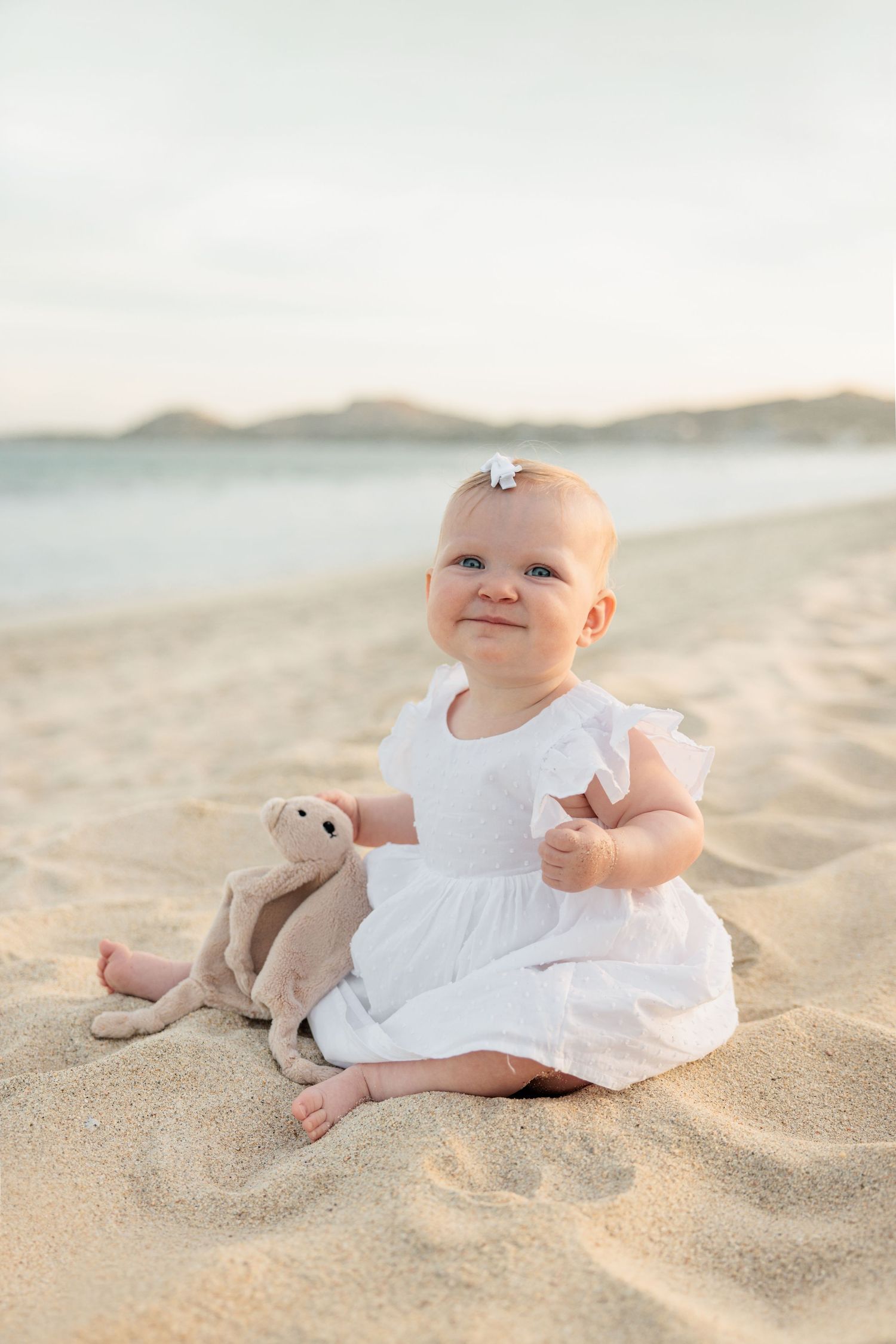 Smiling baby girl in white dress sitting on sandy beach with stuffed animal toy at sunset.