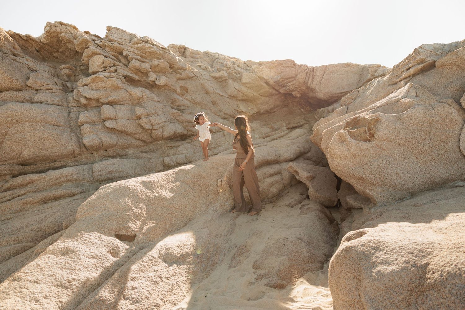 Two women exploring dramatic sandy rock formations in a desert landscape under a bright sky.