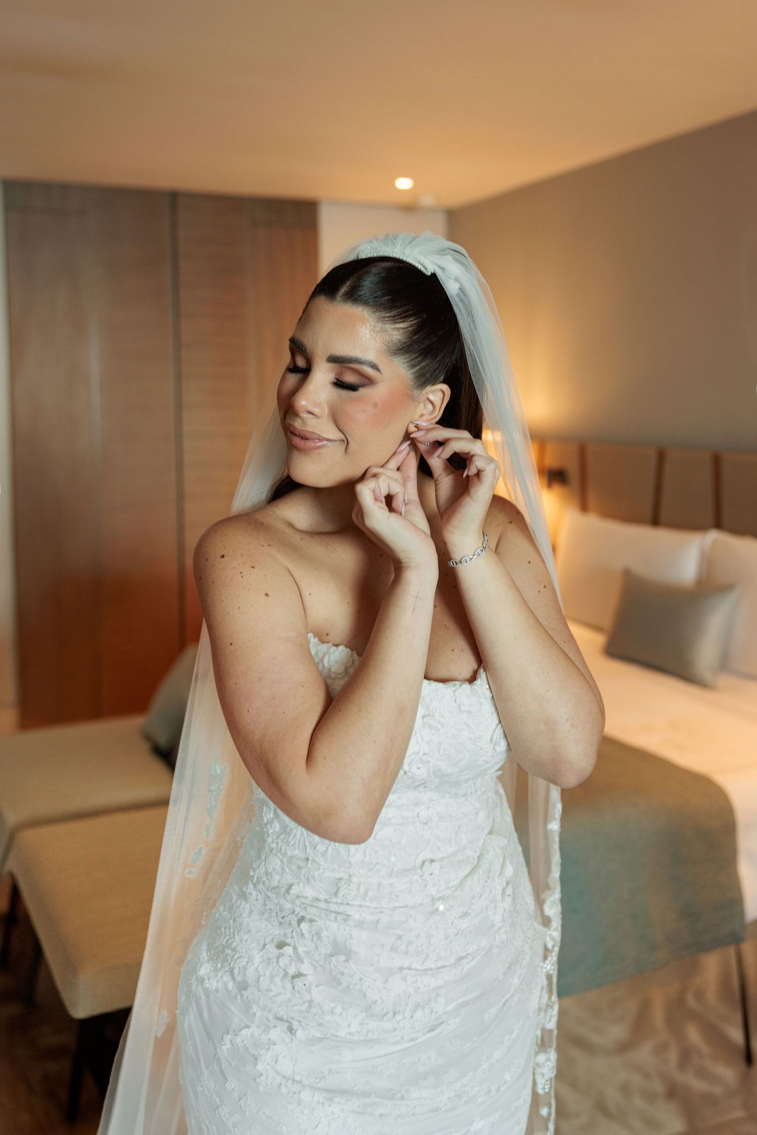 Bride in white lace strapless gown and veil adjusting earrings in a warmly lit hotel room.
