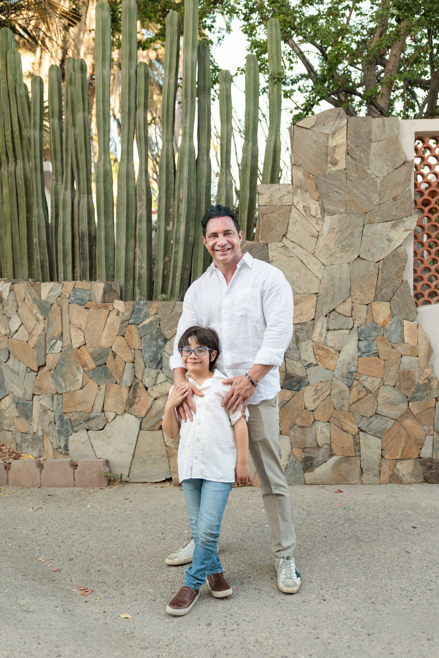 A smiling man in a white shirt poses with a young girl in glasses outdoors near a stone wall and tall cacti.