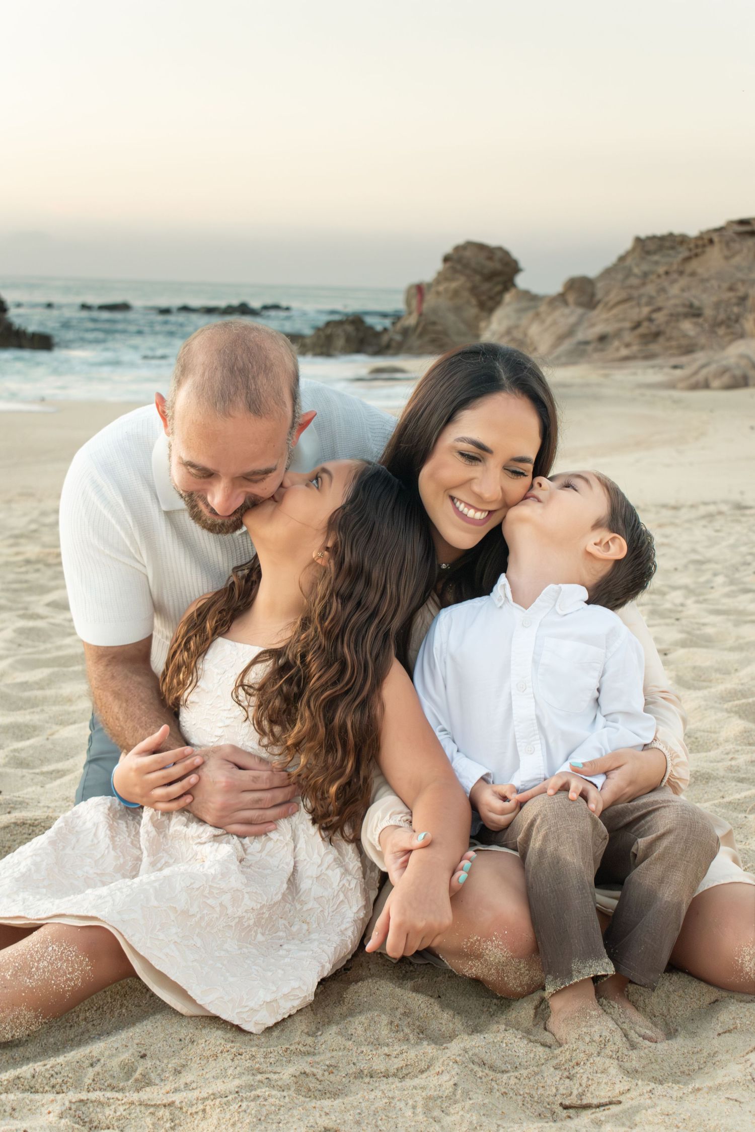 Family of four sharing kisses and hugs while sitting on sandy beach together.