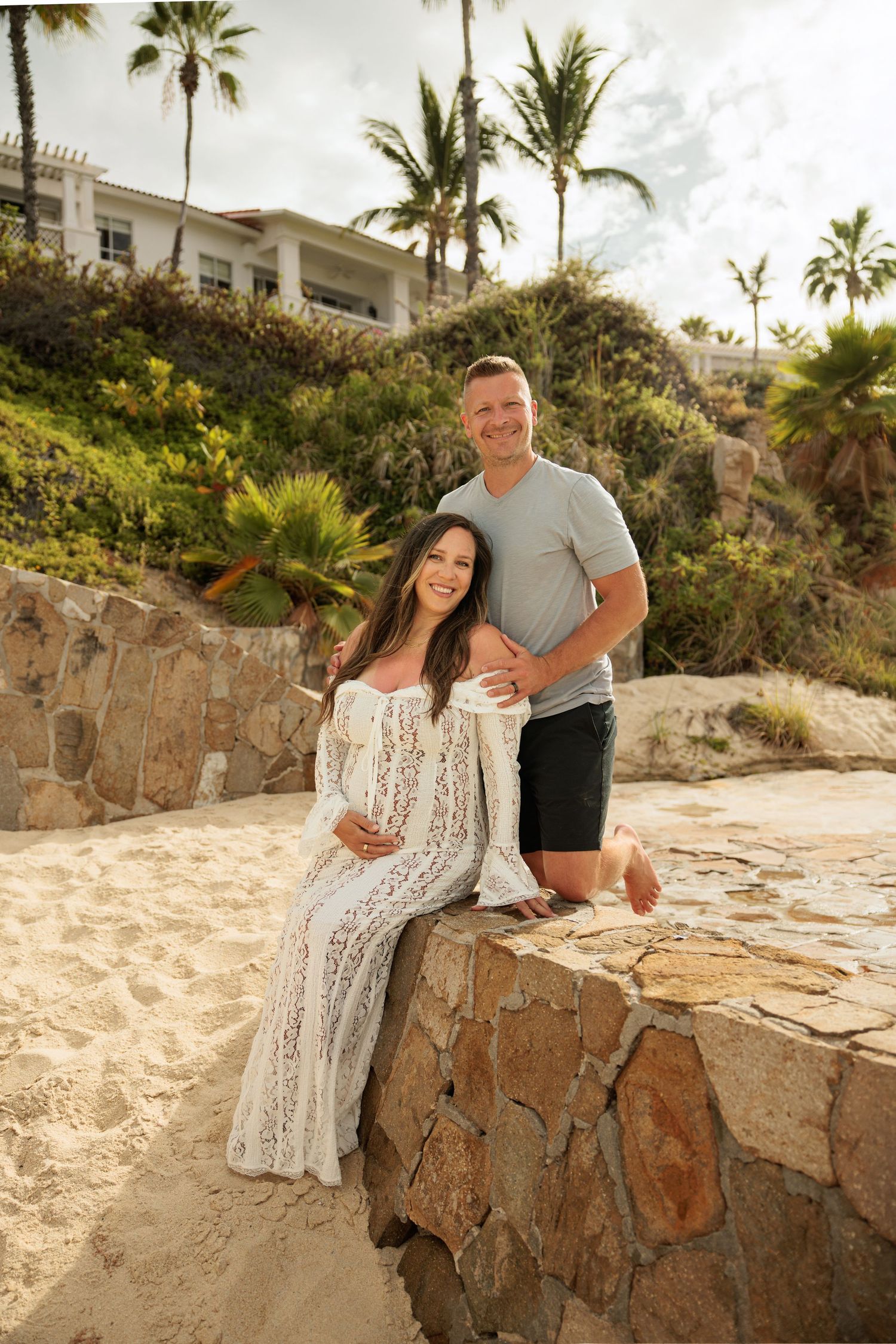 Pregnant woman in white lace dress with partner on sandy beach with palm trees and resort behind.