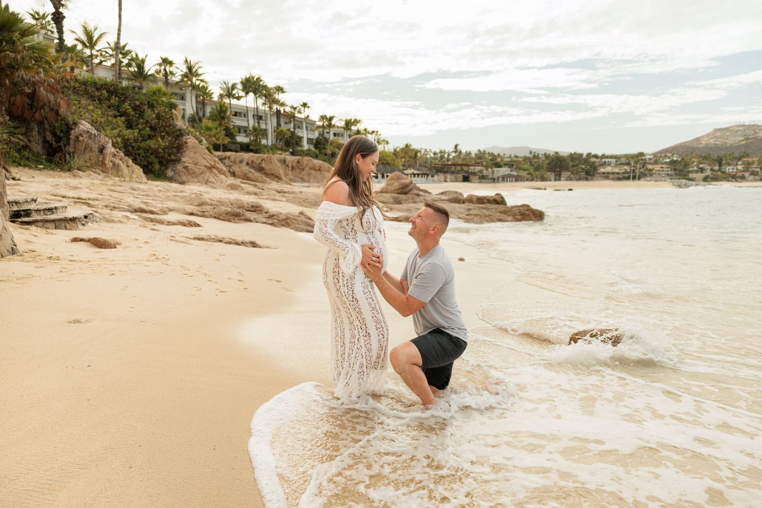 Couple sharing a romantic moment on a sandy Hawaiian beach, woman in white lace dress, waves washing around them.
