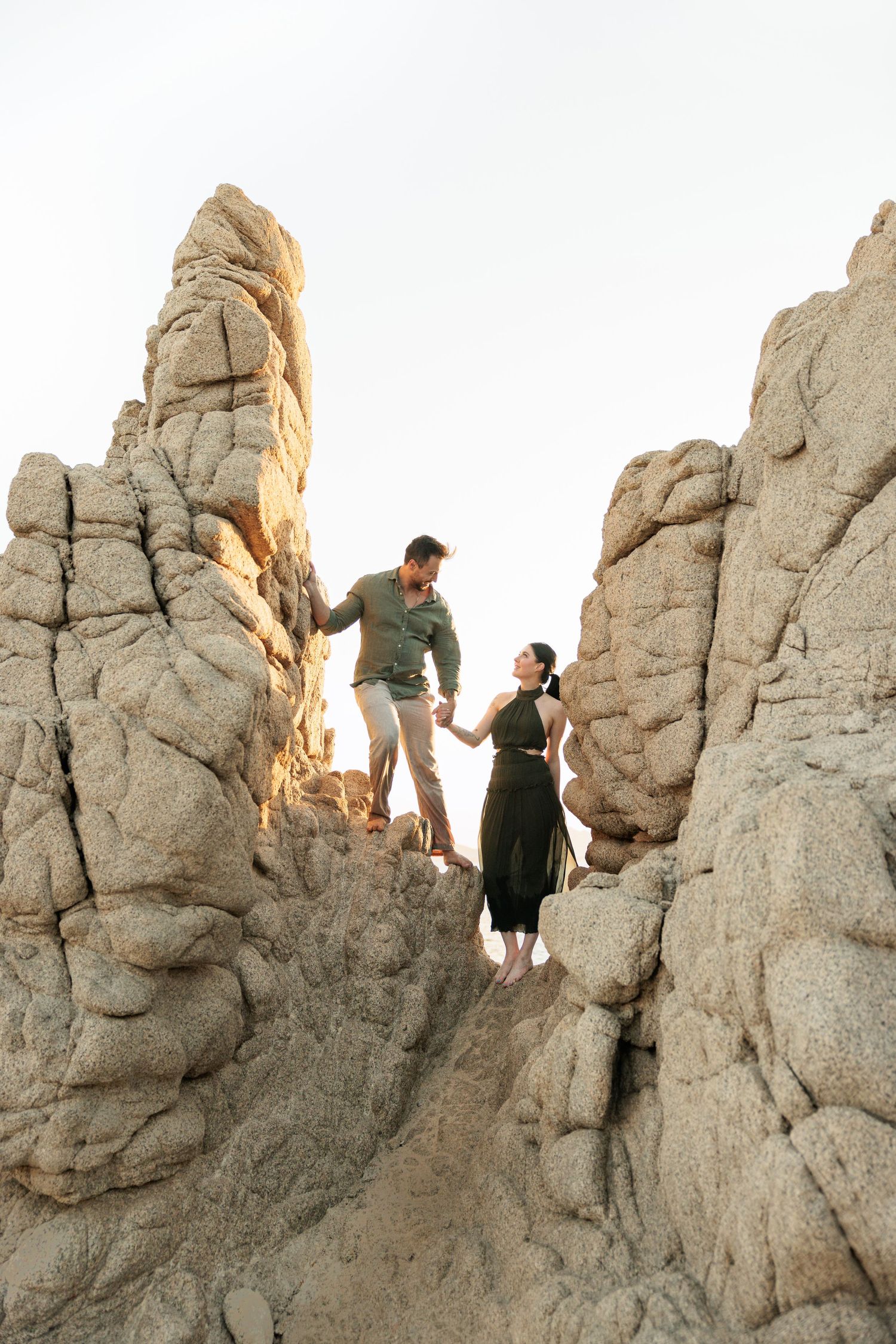 Couple holding hands on rocky coastal formation, man in green shirt and woman in olive dress, standing between tall sandstone rocks.