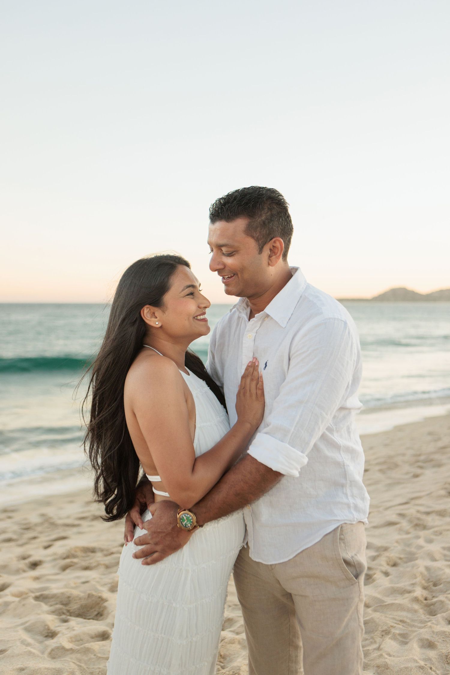 Happy couple embracing on a sandy beach at sunset, woman in white dress laughing with man in white shirt.