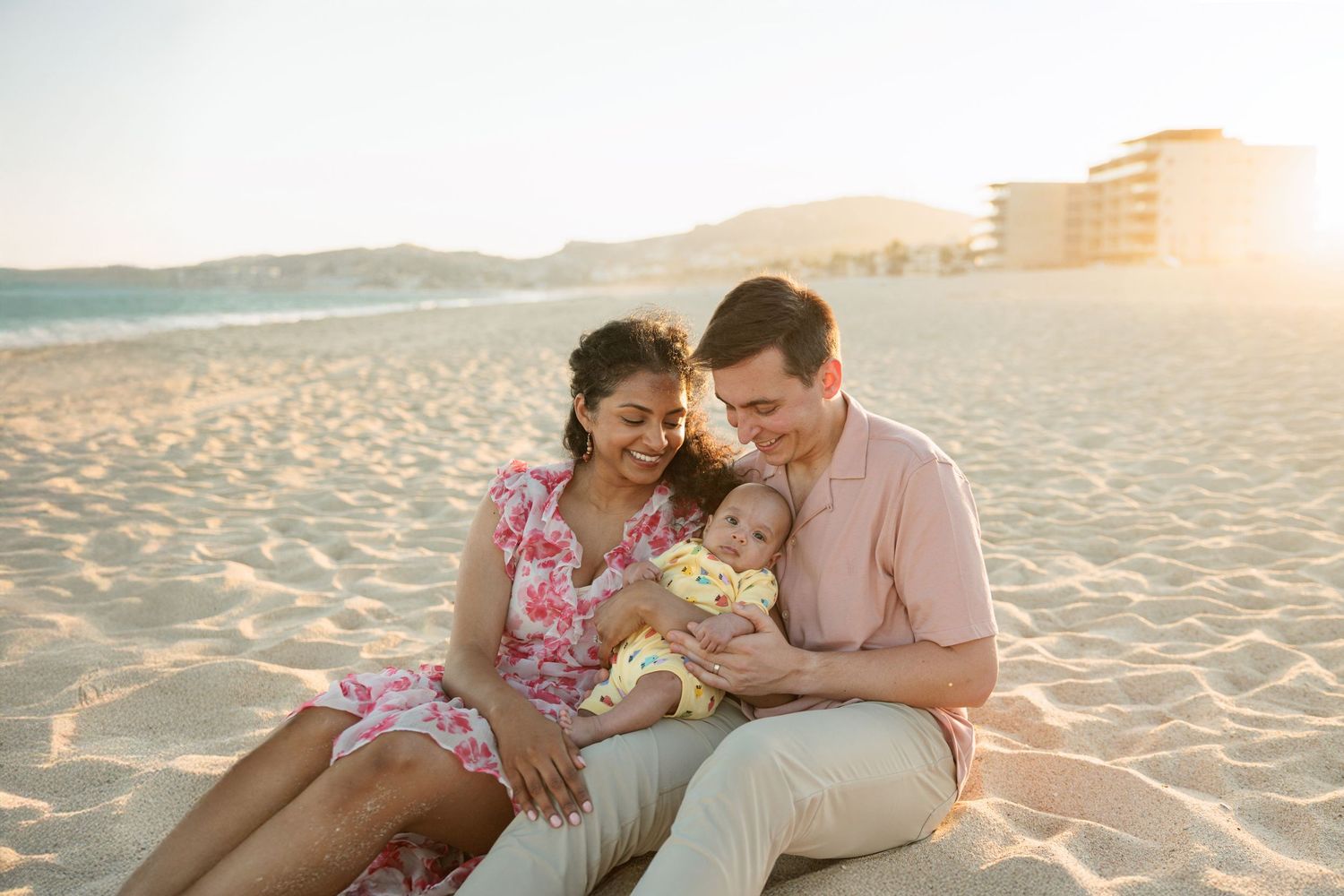 A happy family sits on a sunny beach, parents smiling at their baby in a golden sunset setting.
