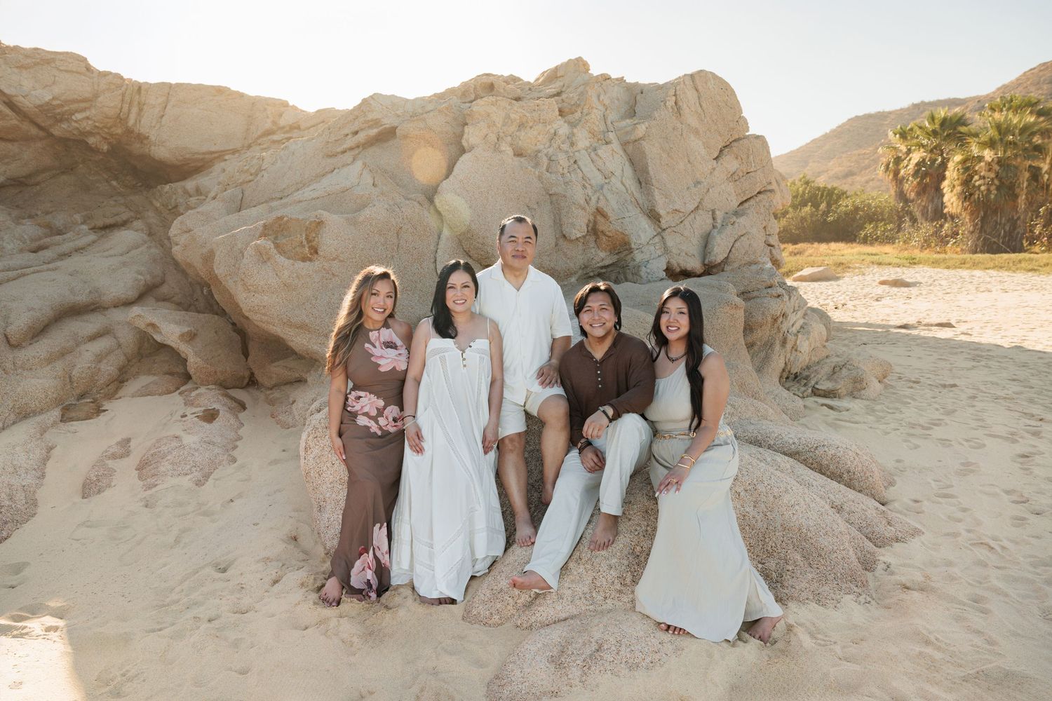 Group of five people posing together on sandy beach with large rock formations in warm golden light.