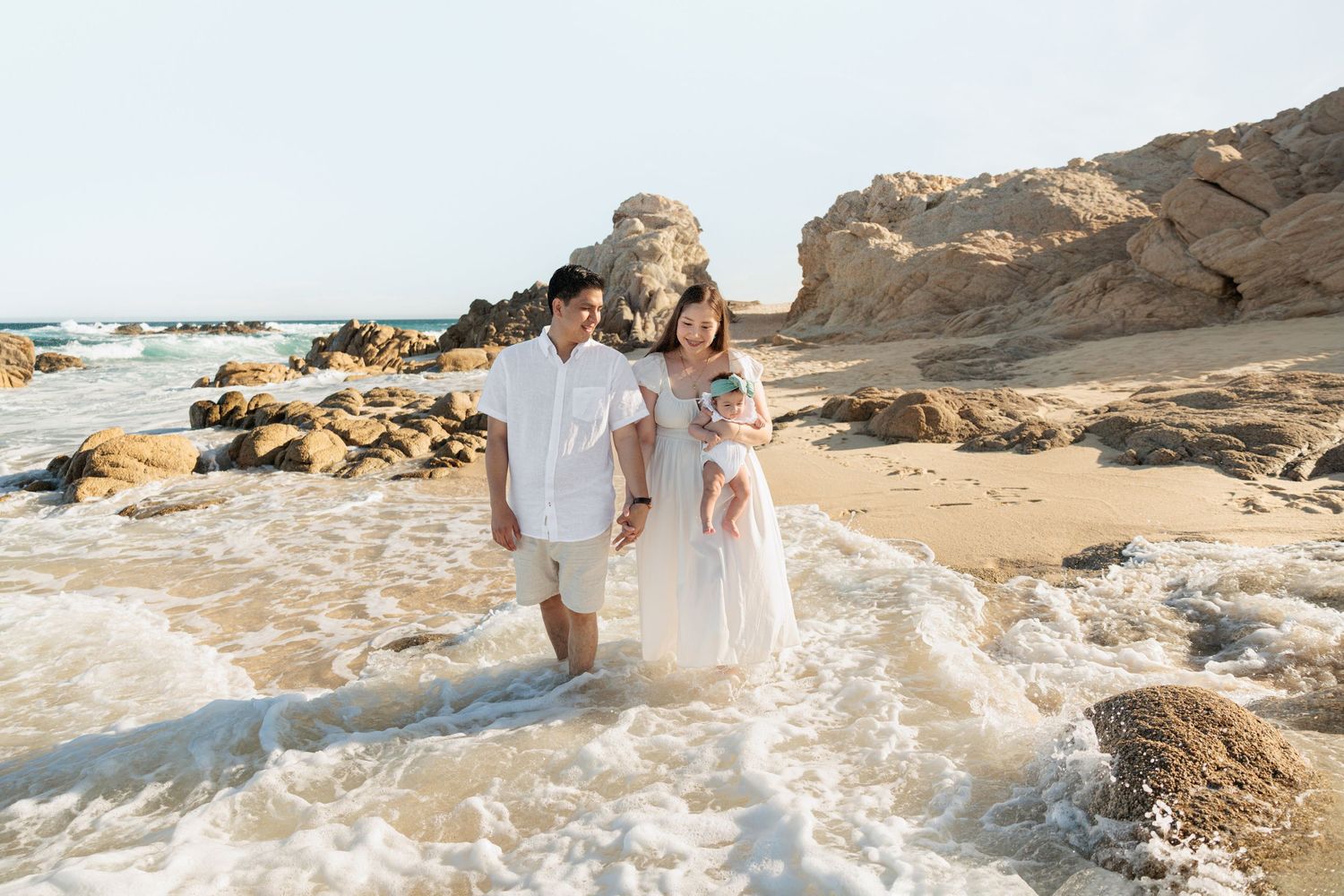 Family of three standing in ocean waves on a sandy beach with rocky cliffs during golden hour.