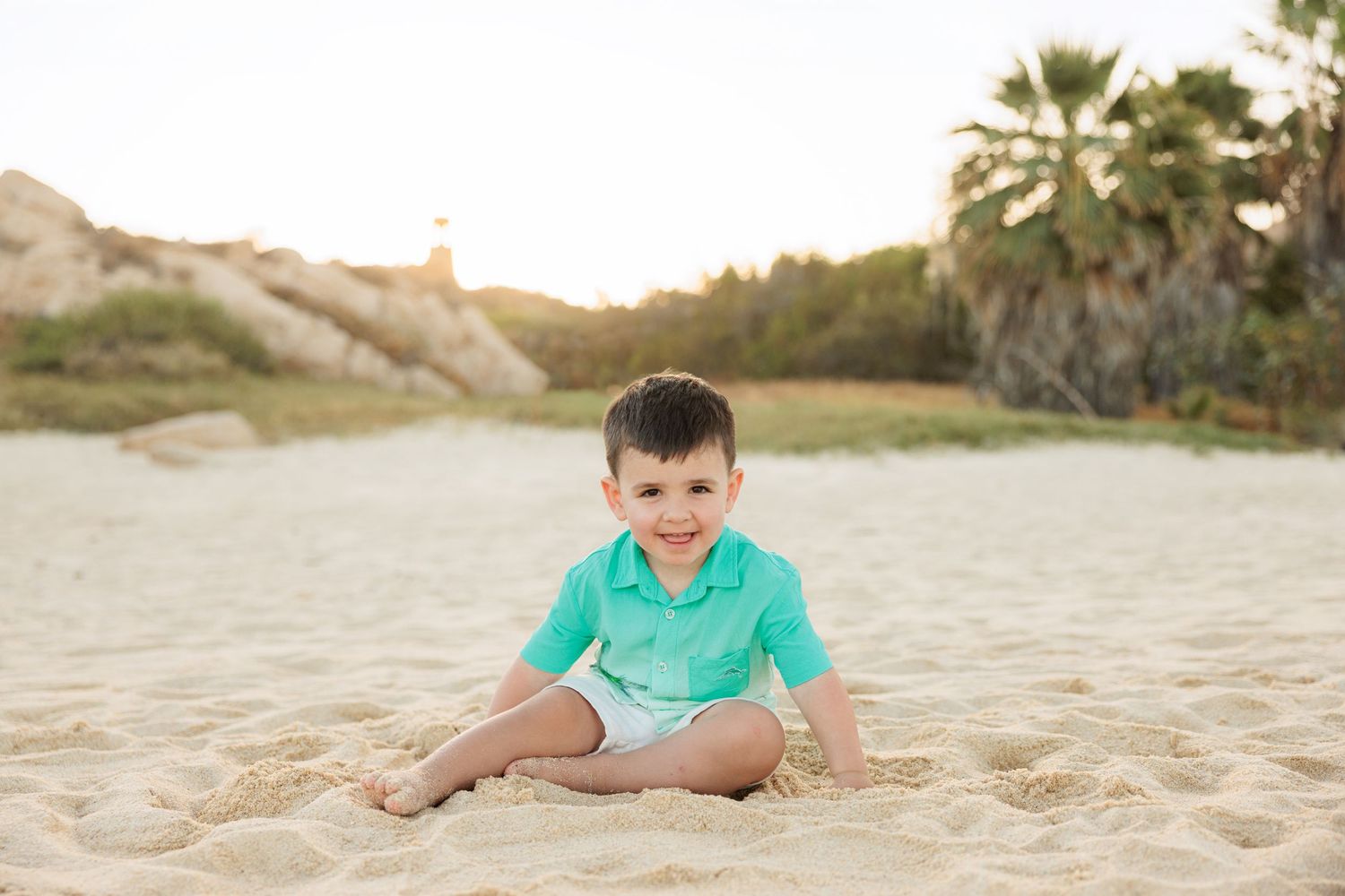 A young boy in a teal shirt and white shorts sits smiling on sandy beach with palm trees and rocks behind him.