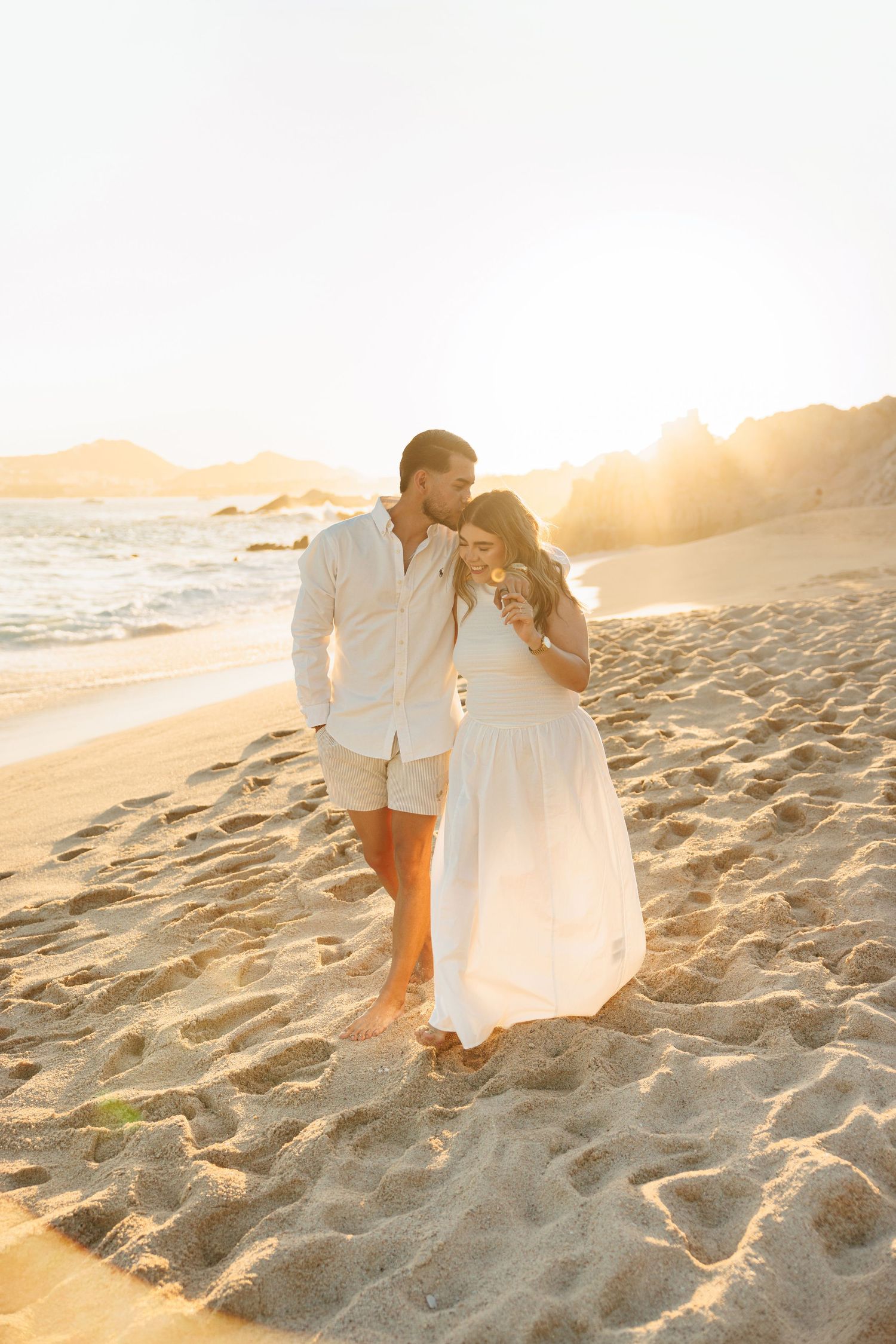 Couple walks on sunny beach at golden hour, man kisses womans head as she wears a flowing white dress.