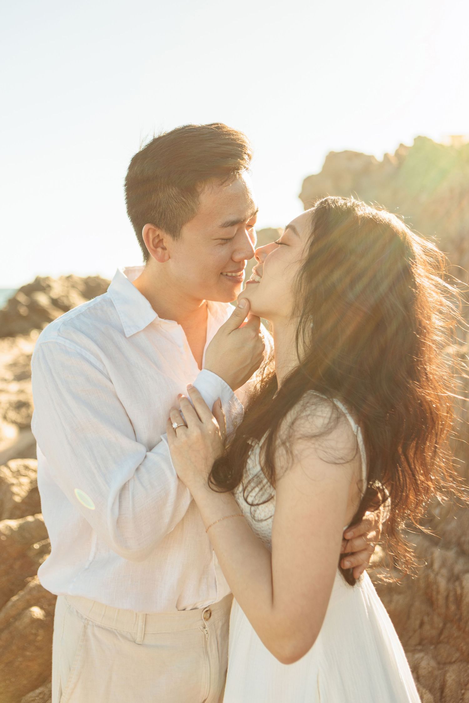 Couple sharing a kiss in golden sunlight on rocky beach, woman in white dress, man in white shirt.