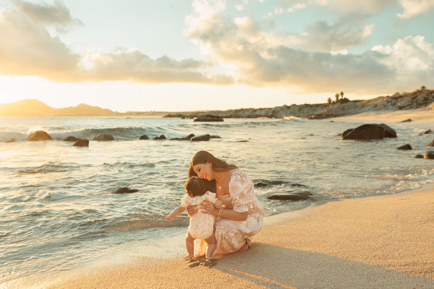 Mother and child share a tender moment on a golden sandy beach at sunset with ocean waves in the background.