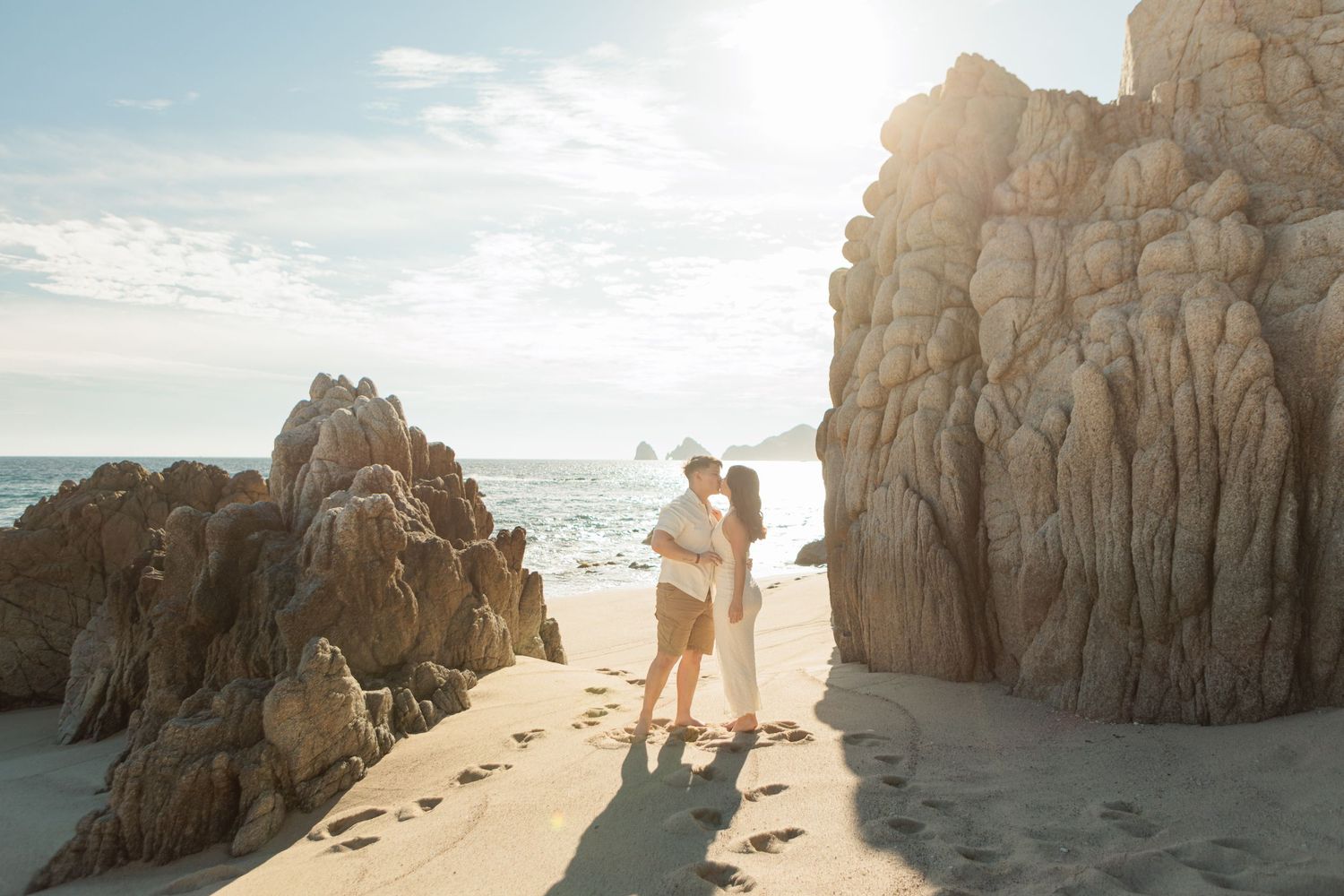 Romantic couple kissing between dramatic rock formations on a sandy beach at sunset.