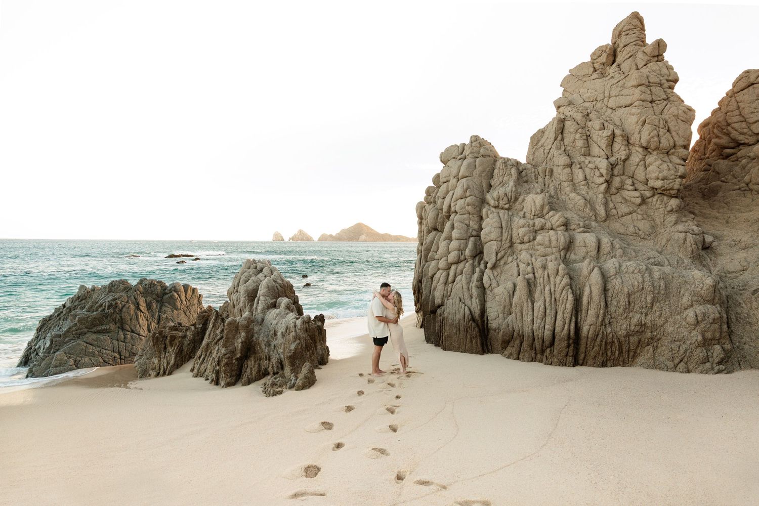 Couple embracing on sandy beach with dramatic rock formations and ocean waves in the background, footprints in sand.
