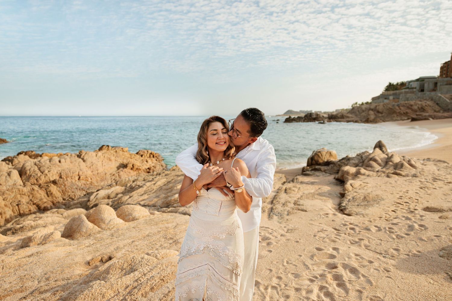 Couple in white outfits embracing on a sandy beach with rocky coastline and blue ocean in the background.