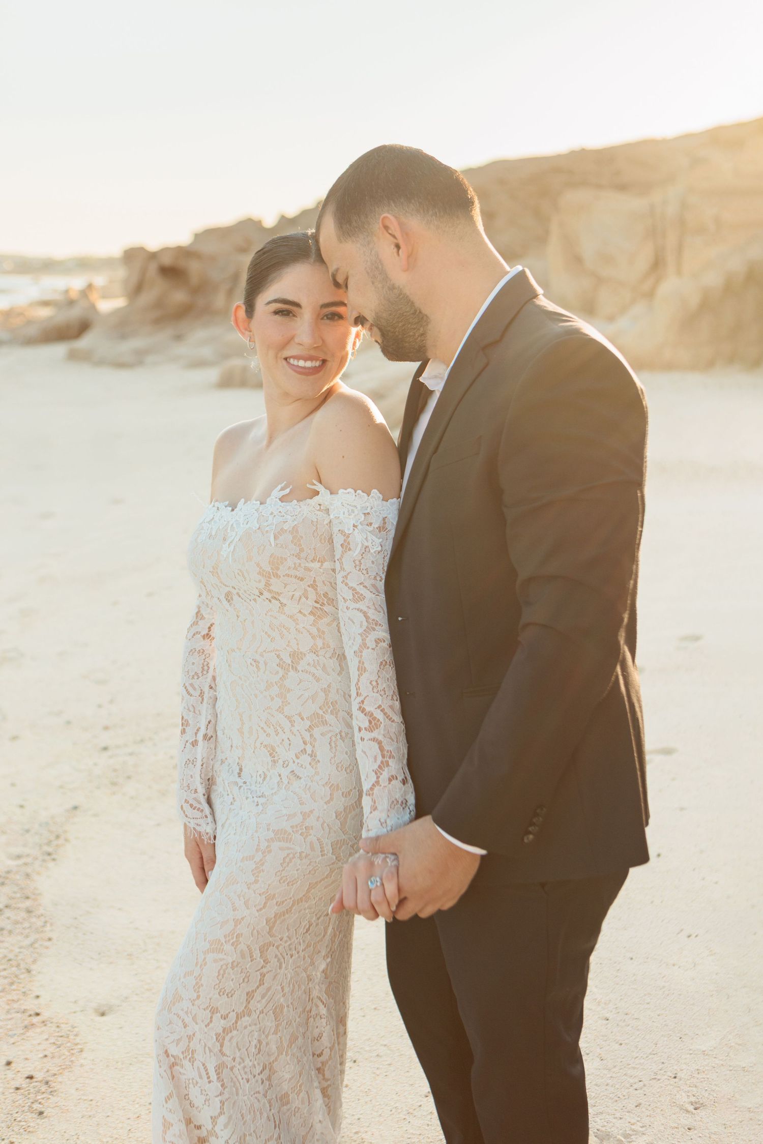 A bride in a lace off-shoulder gown and groom in a black suit hold hands on a sunlit sandy beach.