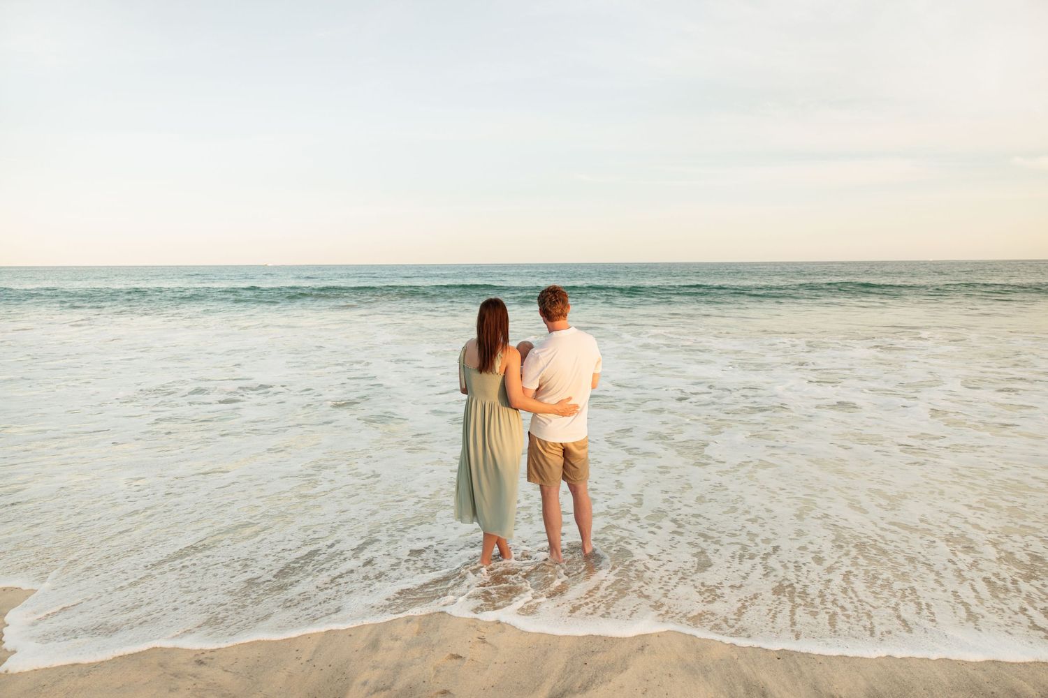 Couple standing together at the ocean's edge on a sandy beach during golden hour sunset.